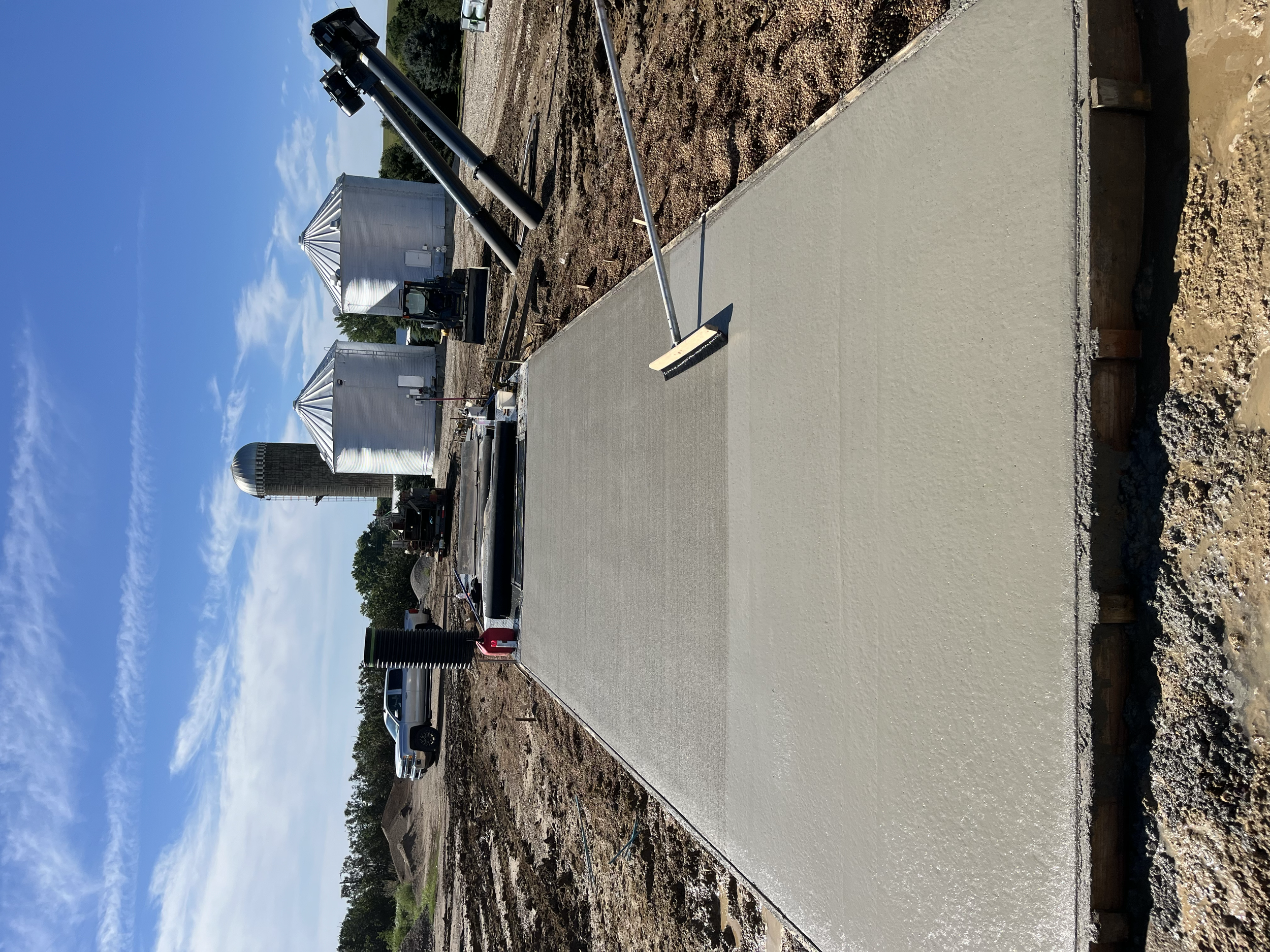 Construction site with freshly poured concrete and construction equipment in the background, partially built road or sidewalk, and a blue sky.