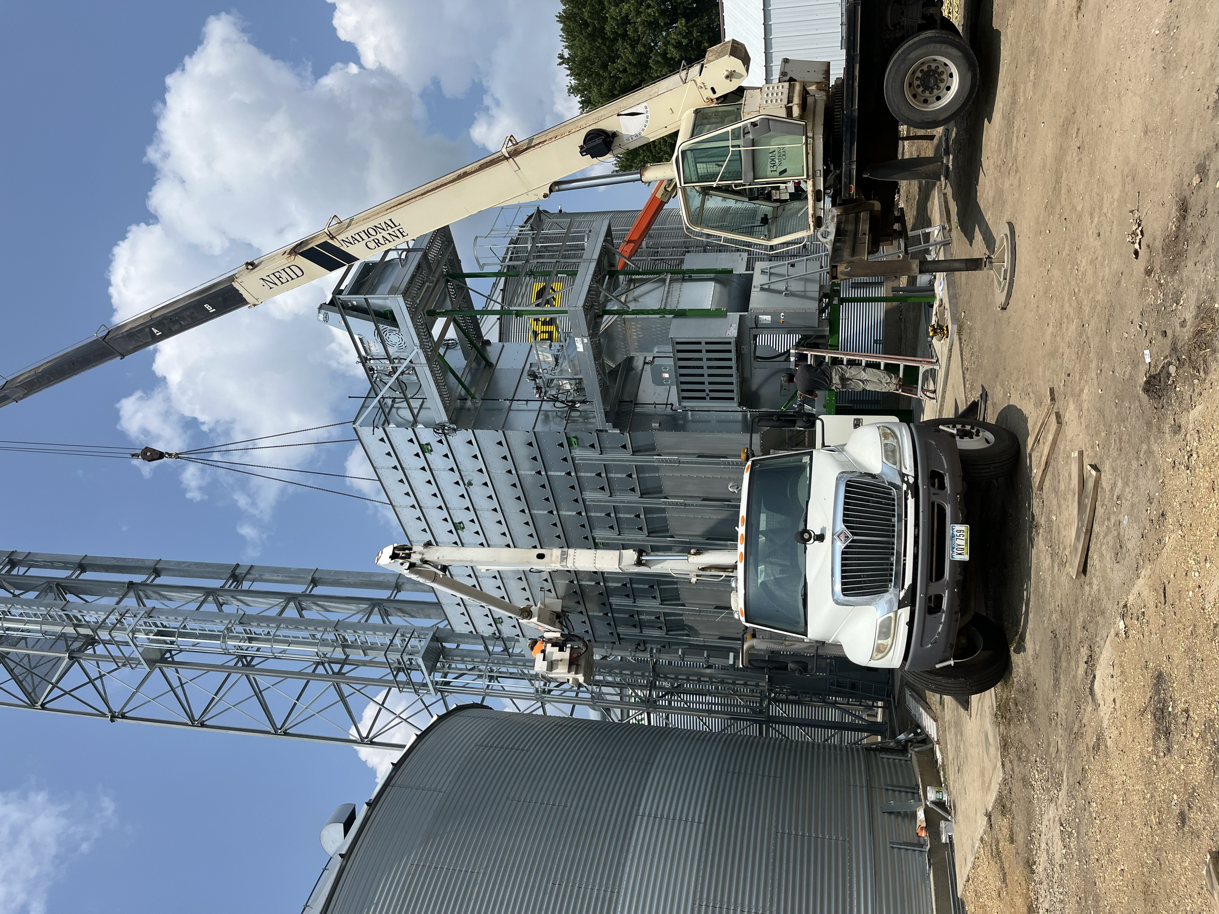 A large industrial crane mounted on a truck is lifting a metal grain bin structure in an outdoor construction site. The sky is partly cloudy, and there are various construction materials and equipment around.