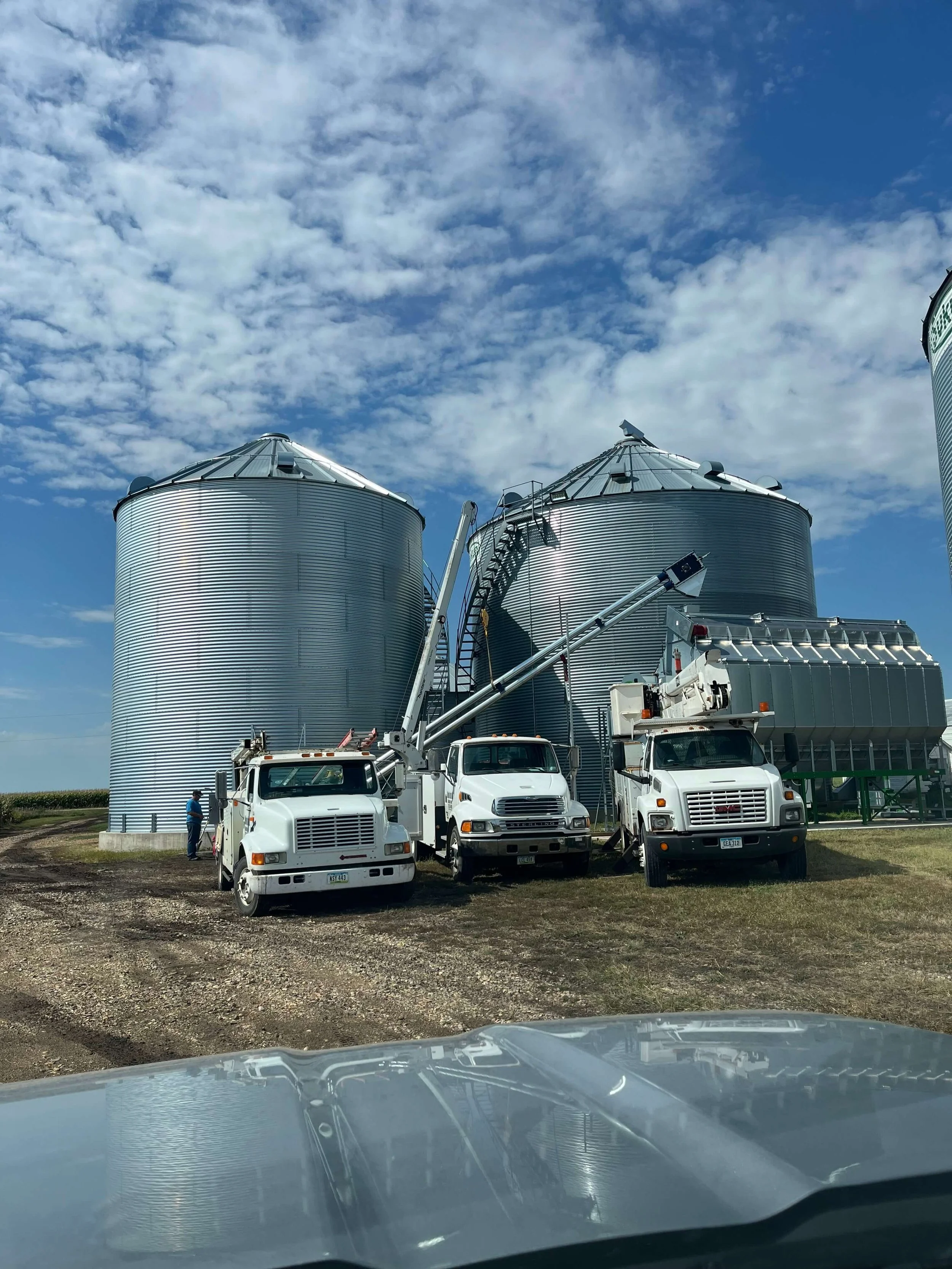Large metal grain silos with trucks parked in front, some workers, and a blue sky with cloud cover.