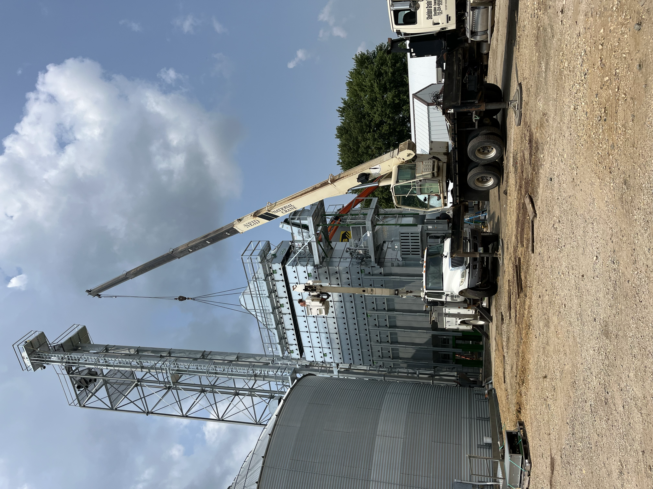 A large tower crane positioned next to blue grain silos on a farm with a white building and green trees in the background. The ground is covered in dirt and gravel.