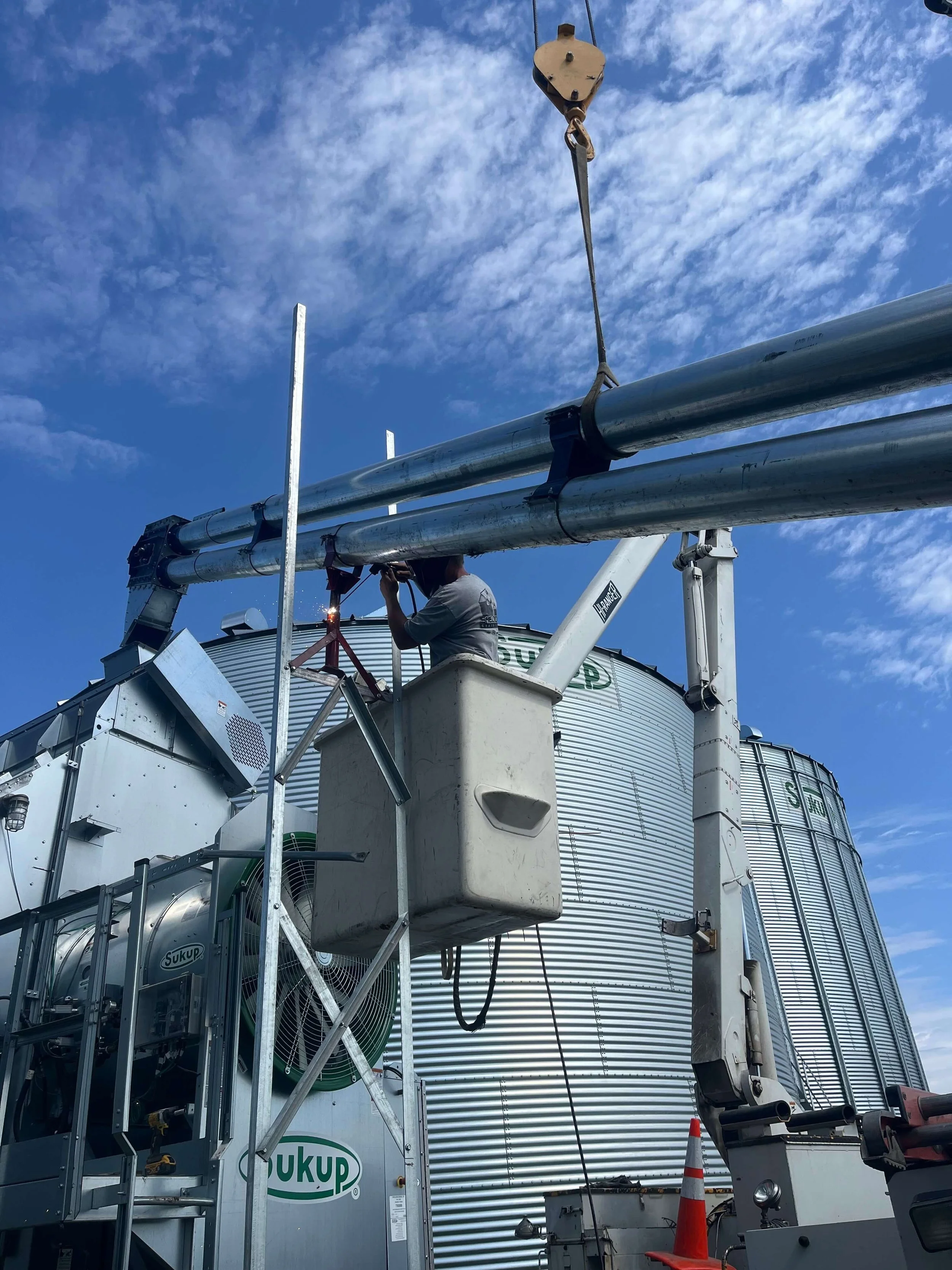 Person working on top of a large industrial grain silo, with metal pipes and equipment, under a blue sky with scattered clouds.