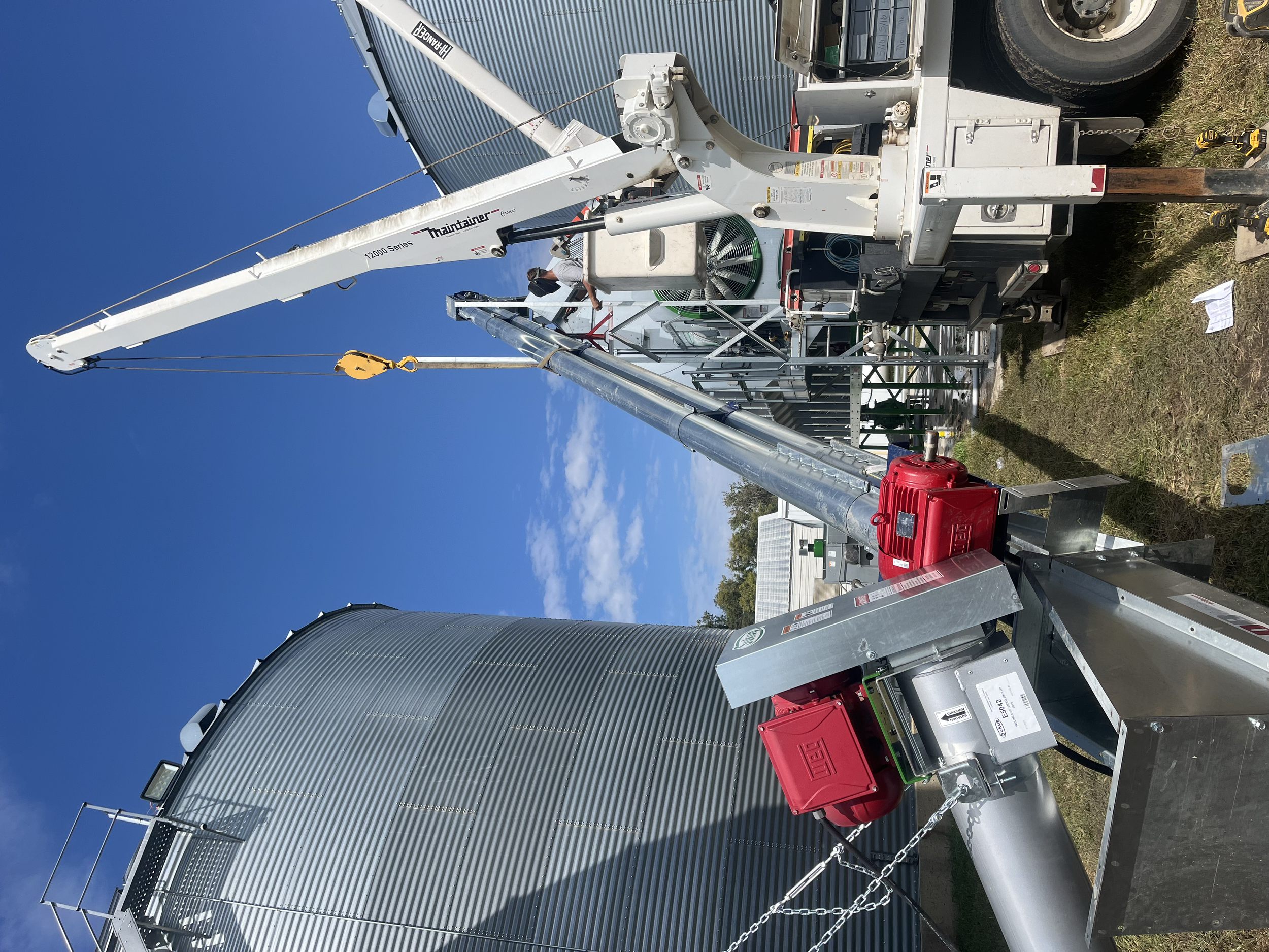 A mobile crane attached to a trailer, with a person working on the crane, in front of a metal industrial building with curved roofs and a clear blue sky.