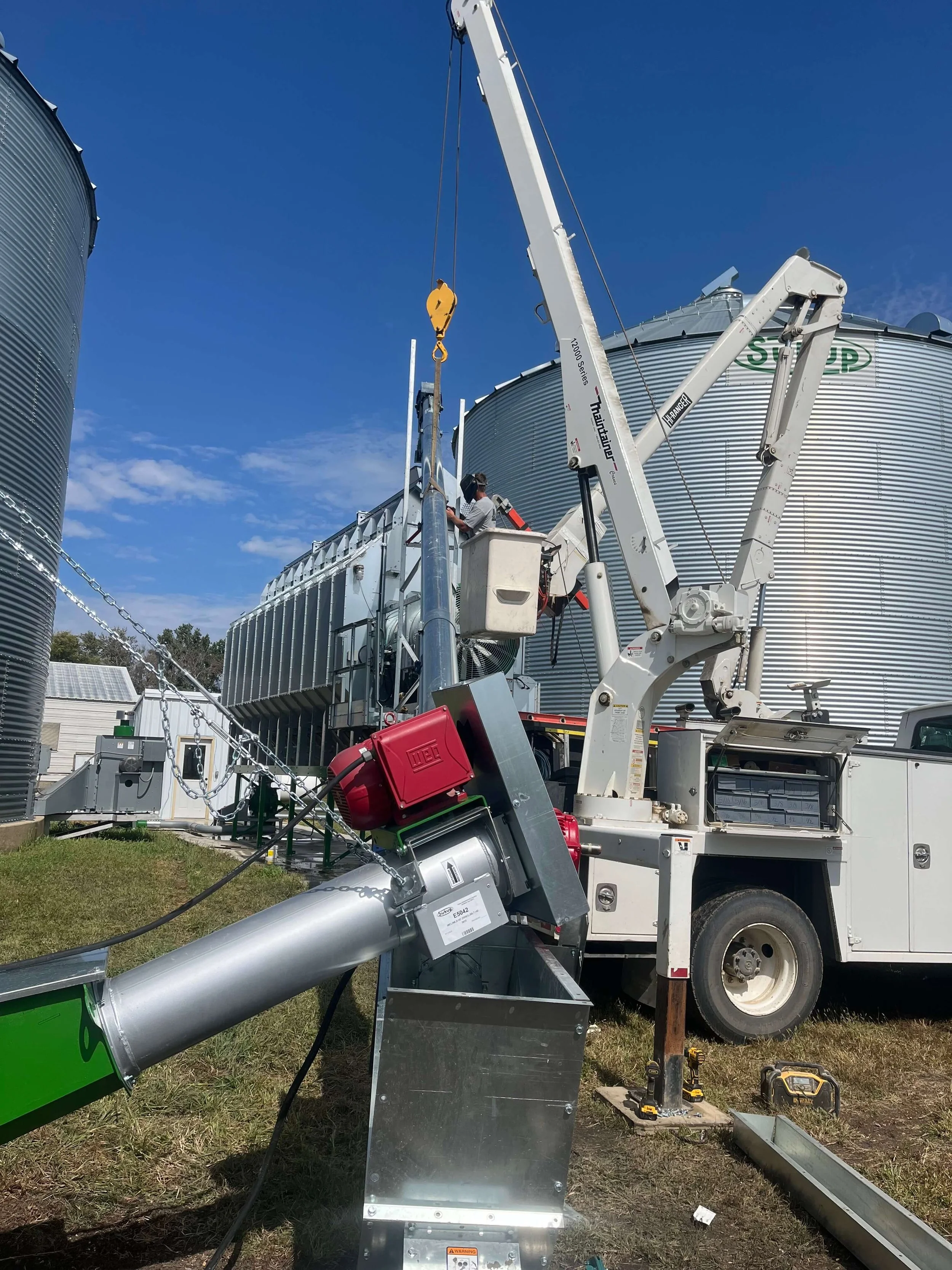 A worker using a boom lift to install or repair a large metal grain silo on a farm. There is other farm equipment and silos visible in the background under a clear blue sky.