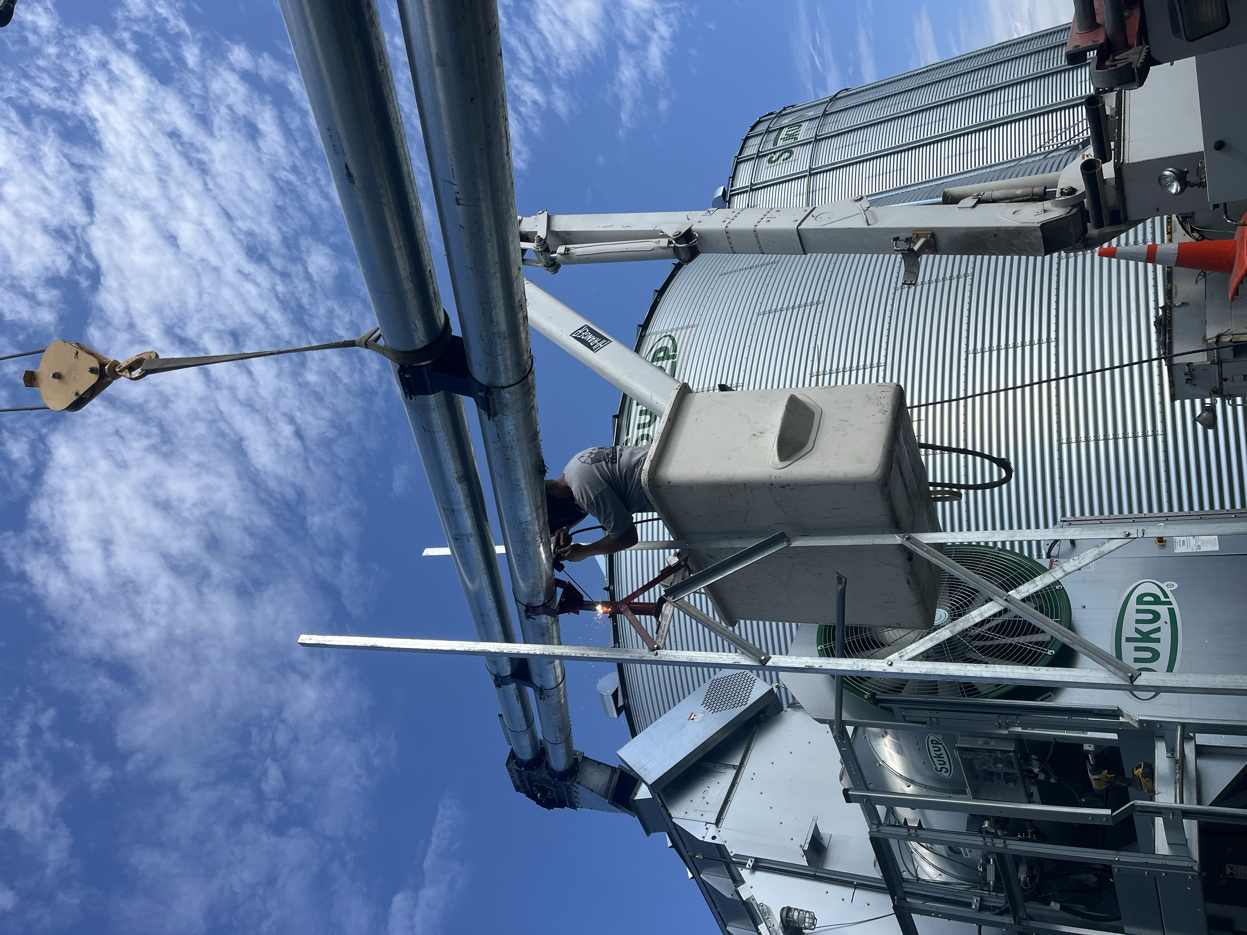 Photo of agricultural equipment, specifically grain silos with metal piping and scaffolding, against a blue sky with scattered white clouds.