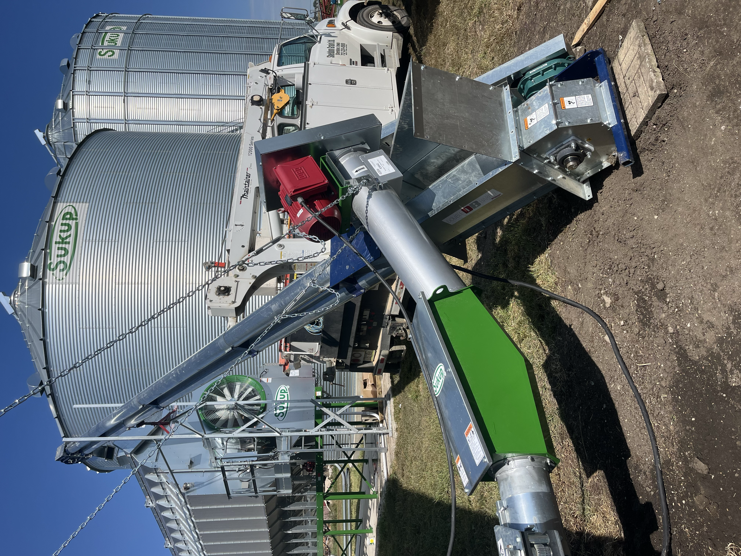 A large grain storage silo with the brand name 'Superior' on it, connected to a mobile grain filling machine with various pipes and machinery, set on a grassy and dirt area under a clear blue sky.
