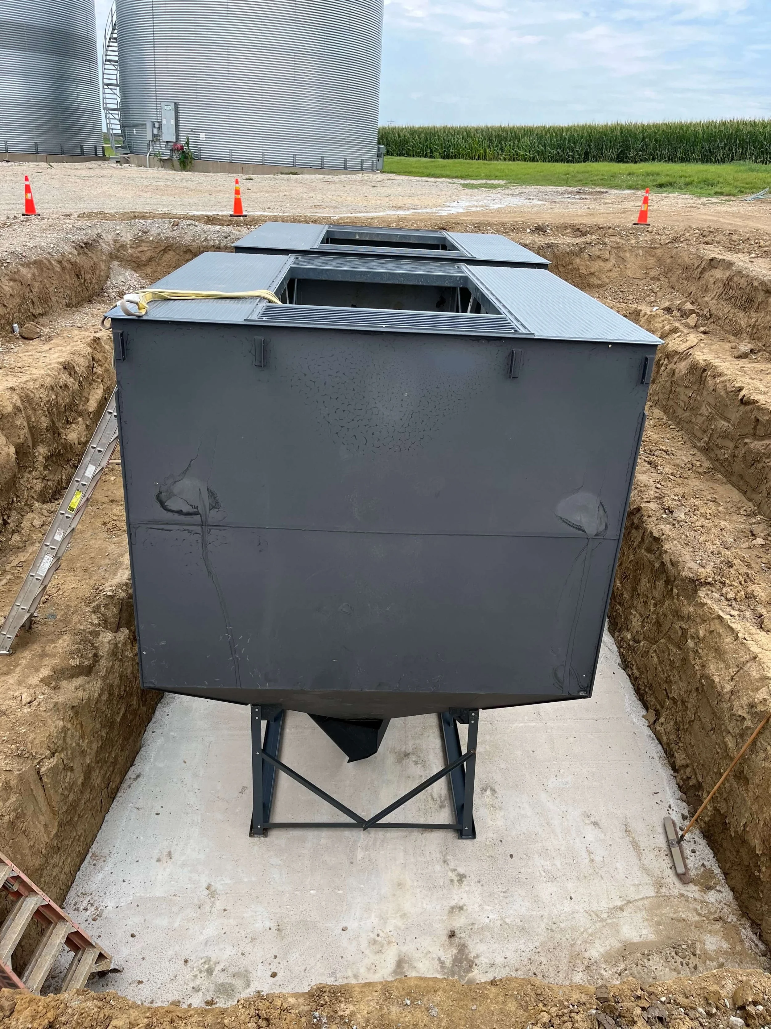 Underground utility vault with a metal cover open, located in a construction site with dirt and gravel surrounding it, with grain silos and farmland in the background.
