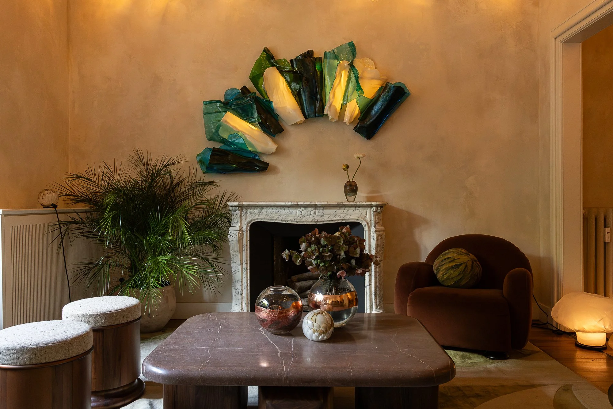 Living room with a marble fireplace, green and black art installation on the wall, potted plants, a brown armchair with a pillow, and decorative vases on a coffee table.