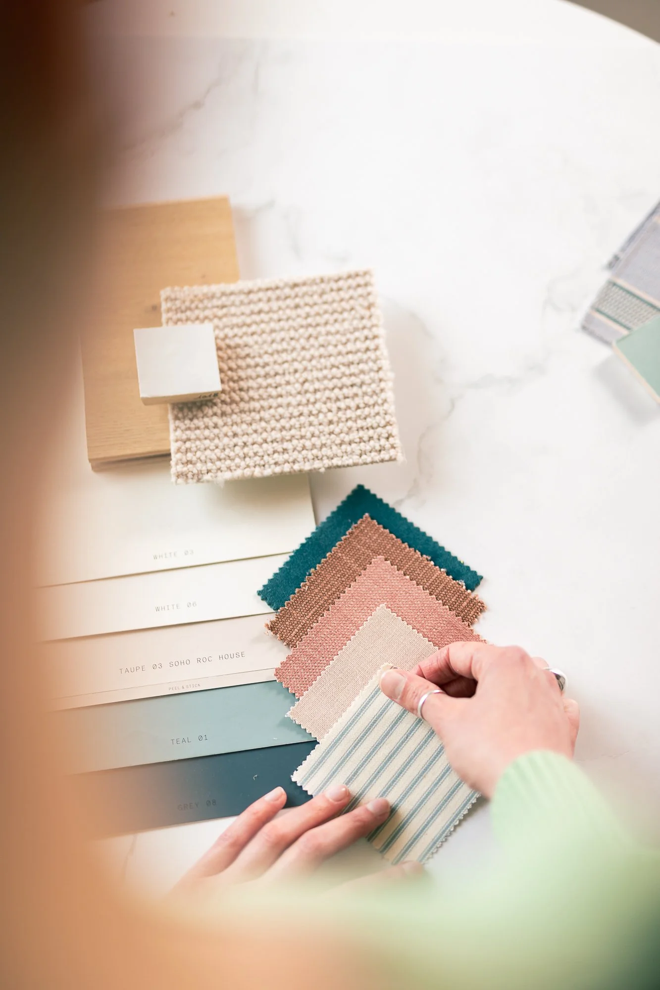 Hands holding fabric swatches and color sample sheets on a table, with material sample cards and color swatches arranged nearby, suggestive of interior design or upholstery planning.
