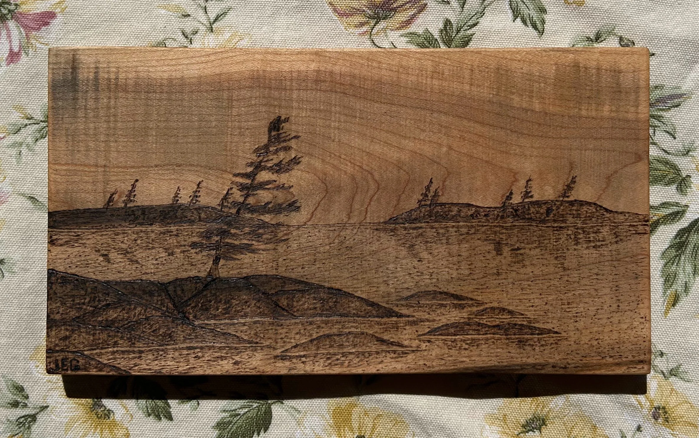 A wooden box featuring a landscape engraving of trees on hilly terrain, placed on a floral-patterned fabric surface.