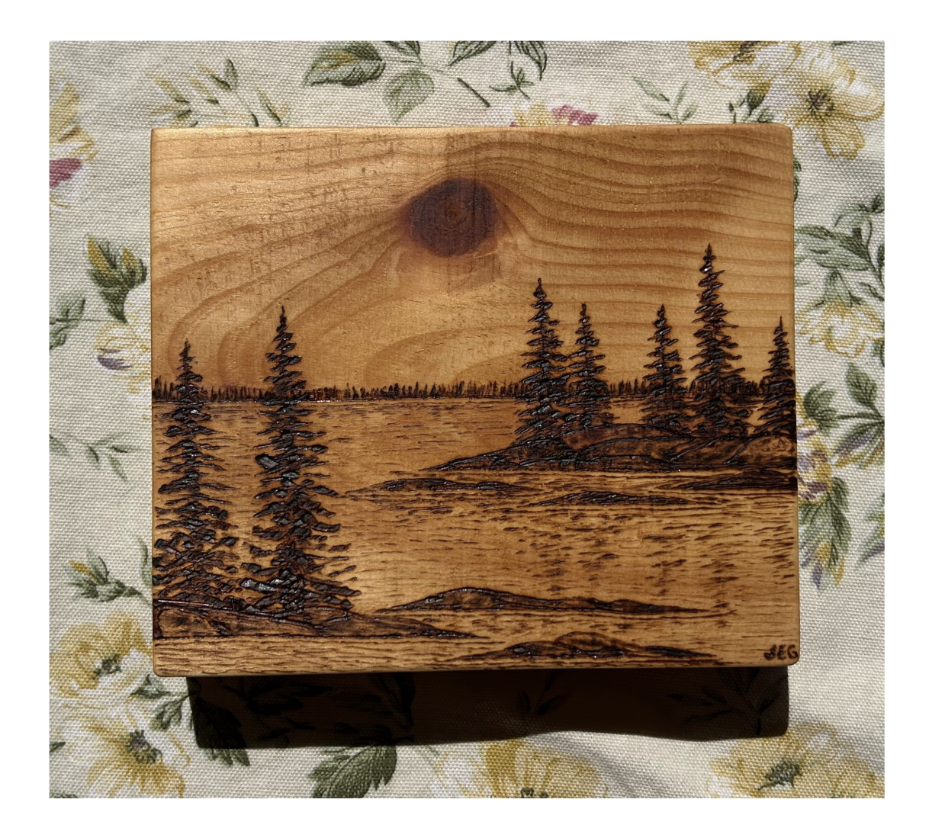 Wooden box with a landscape engraved on its surface, featuring trees, water, and a sky with a moon or sun, set on floral fabric.