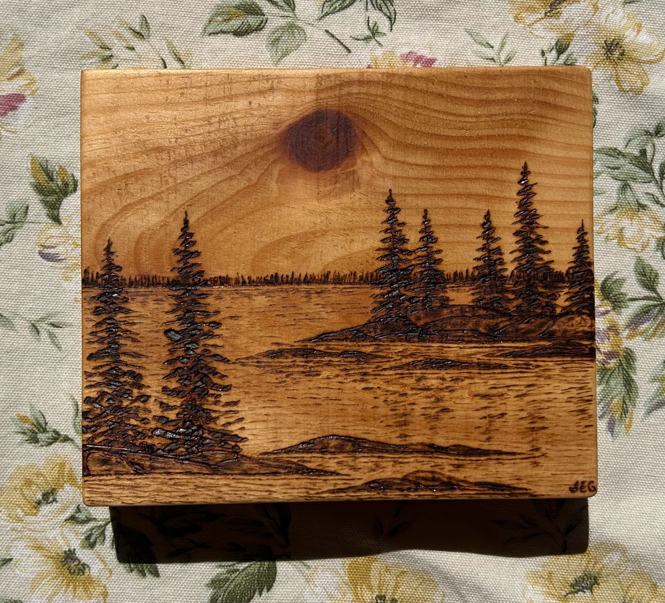 A wooden piece with a landscape laser engraved on it, featuring a large central tree and smaller trees in the background, on a floral patterned tablecloth.