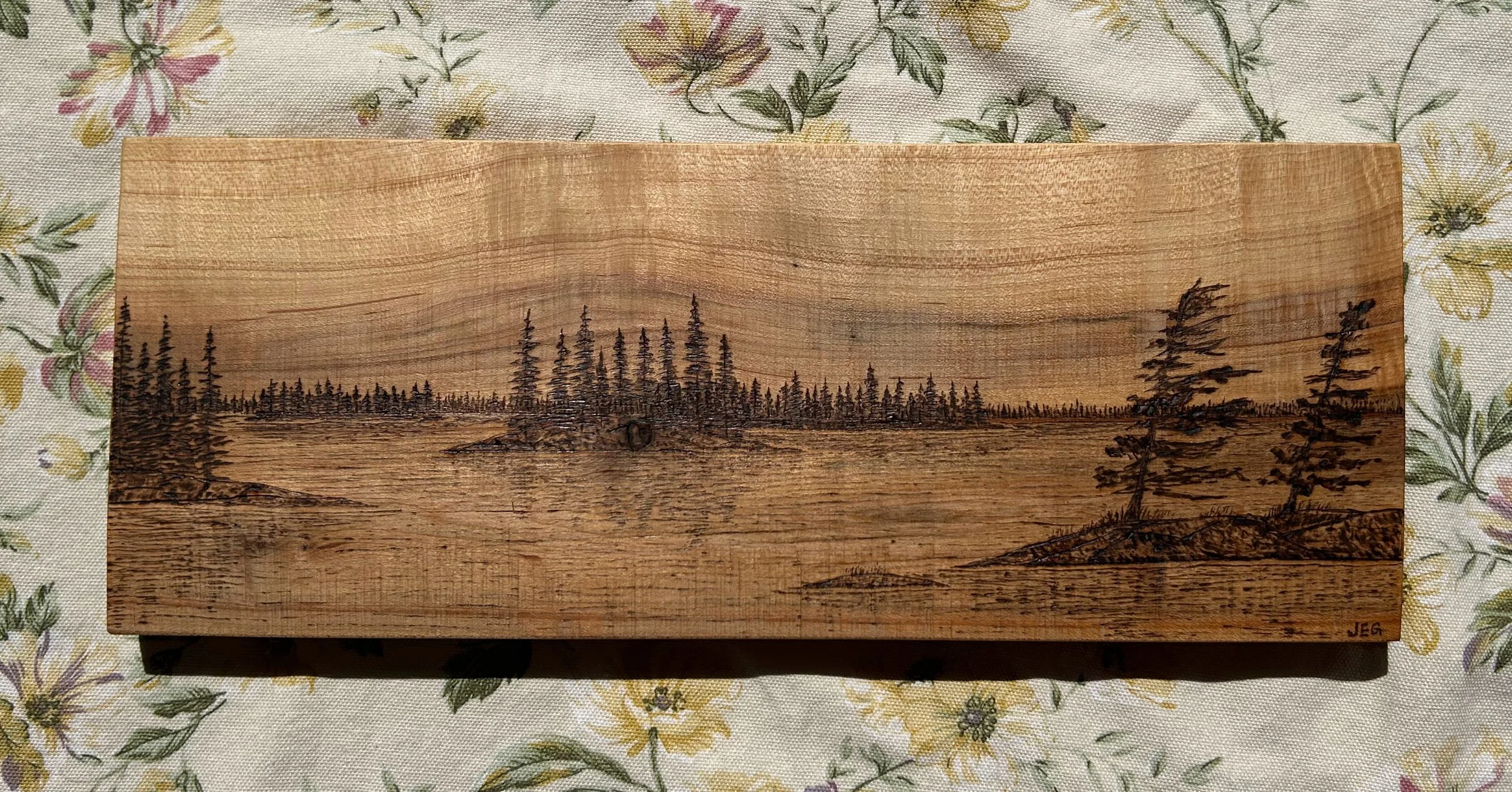 A wooden plaque with a black-engraved landscape scene of a forest and water with trees, etched into the wood, on a floral fabric background.