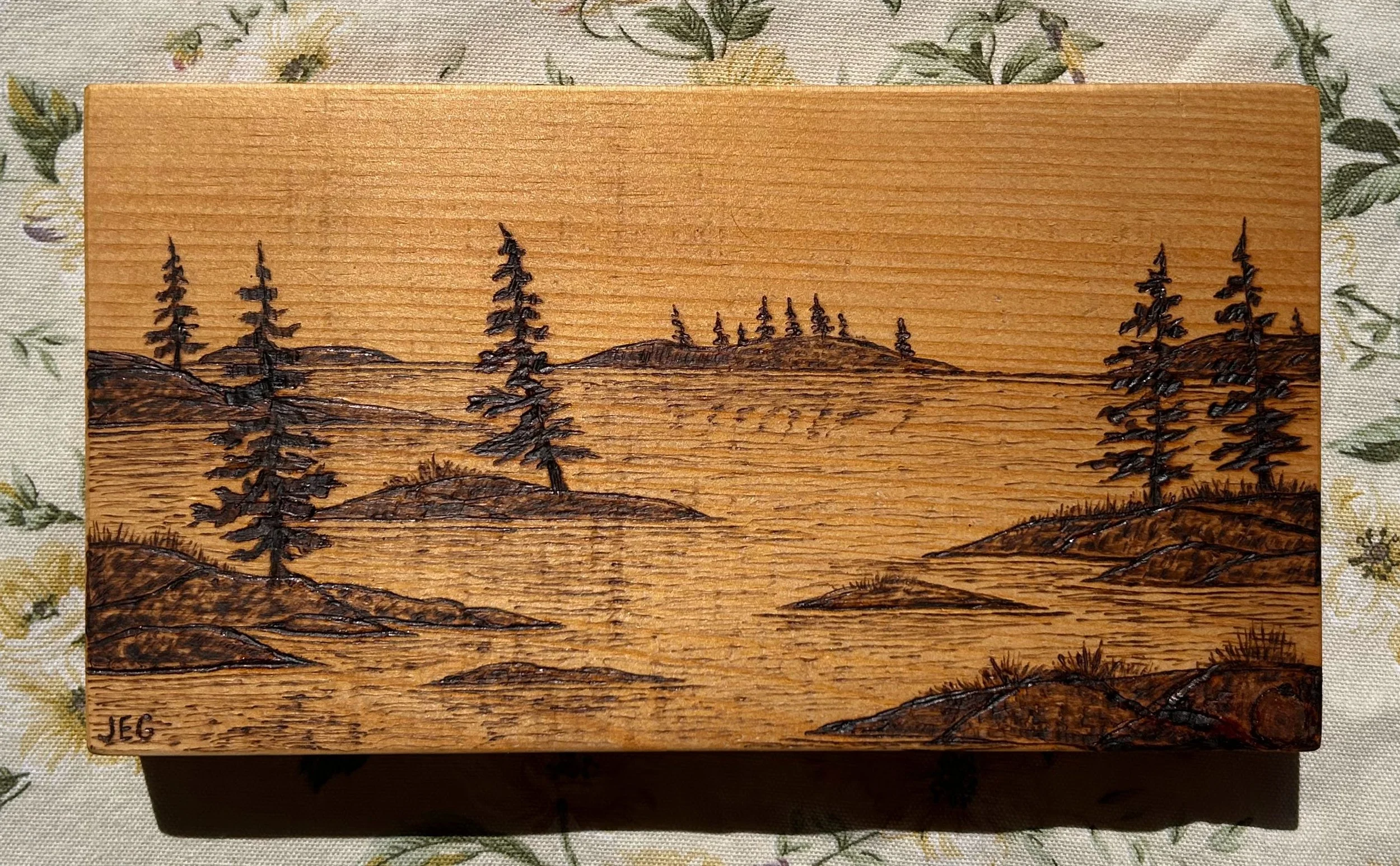 A wooden box with a landscape engraving of trees and hills, signed with initials 'JEG' in the bottom left corner, on a floral-patterned fabric surface.