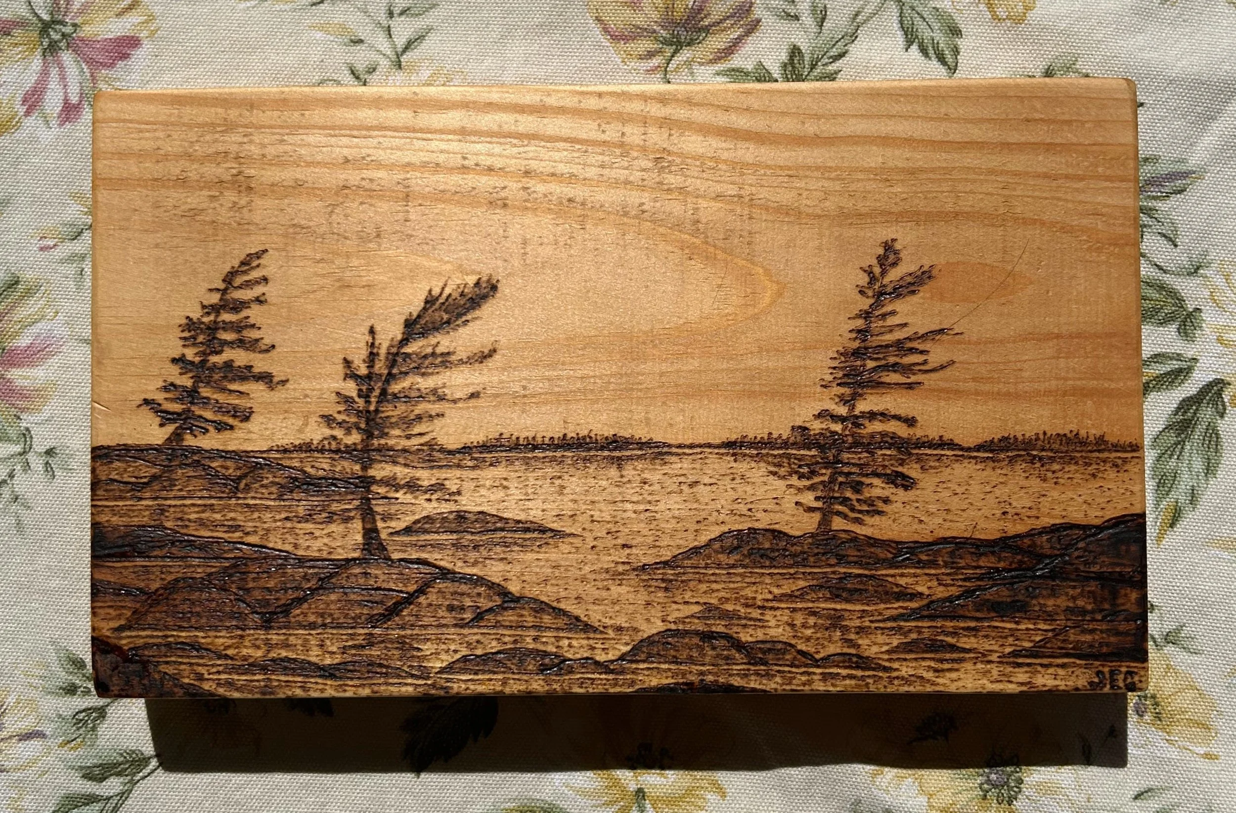 A wooden block with a landscape scene burned into it, depicting two trees, mountains, and ground, placed on a floral tablecloth.
