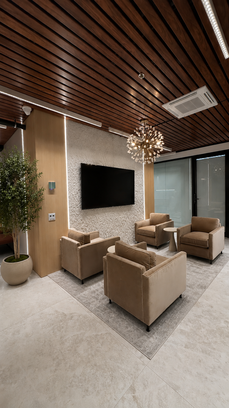 A modern lounge area with beige armchairs, a wall-mounted flat-screen TV, a potted plant, and a decorative chandelier on a textured wall backdrop. Large glass doors are visible on the right.