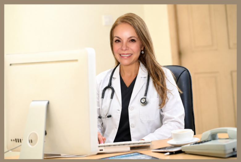 A smiling female doctor sitting at her desk in an office with a computer, stethoscope around her neck, and a cup of coffee.