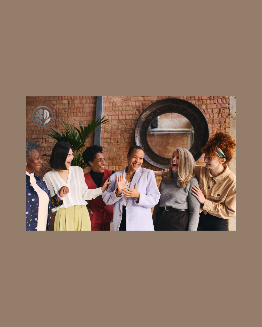 A diverse group of six women smiling and laughing together in an indoor space with a brick wall and a large round mirror behind them.