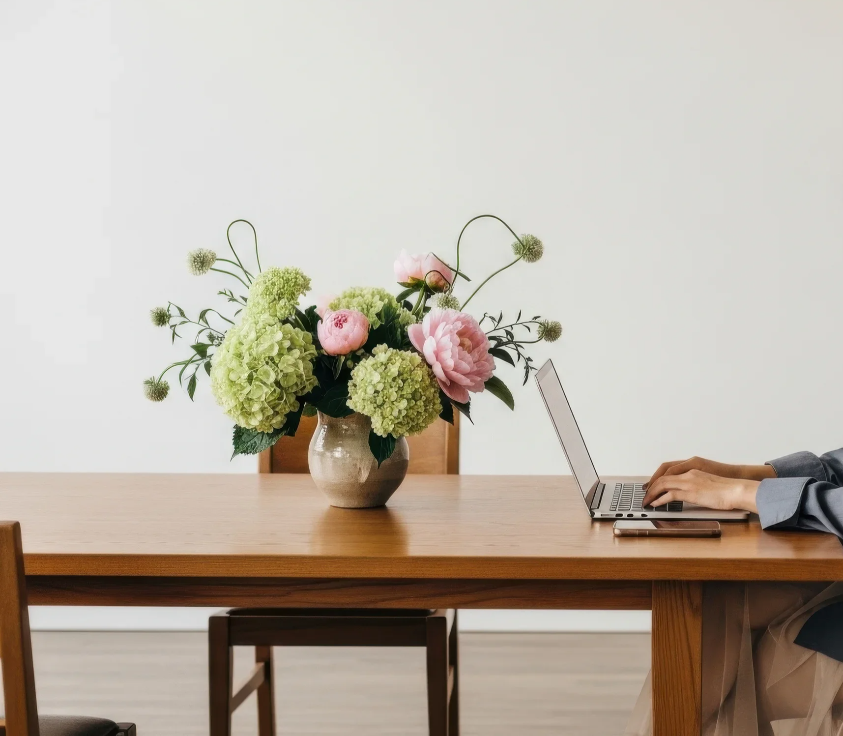 A person typing on a laptop at a wooden dining table with a vase of pink and green flowers, including hydrangeas and peonies, on the left side of the table.