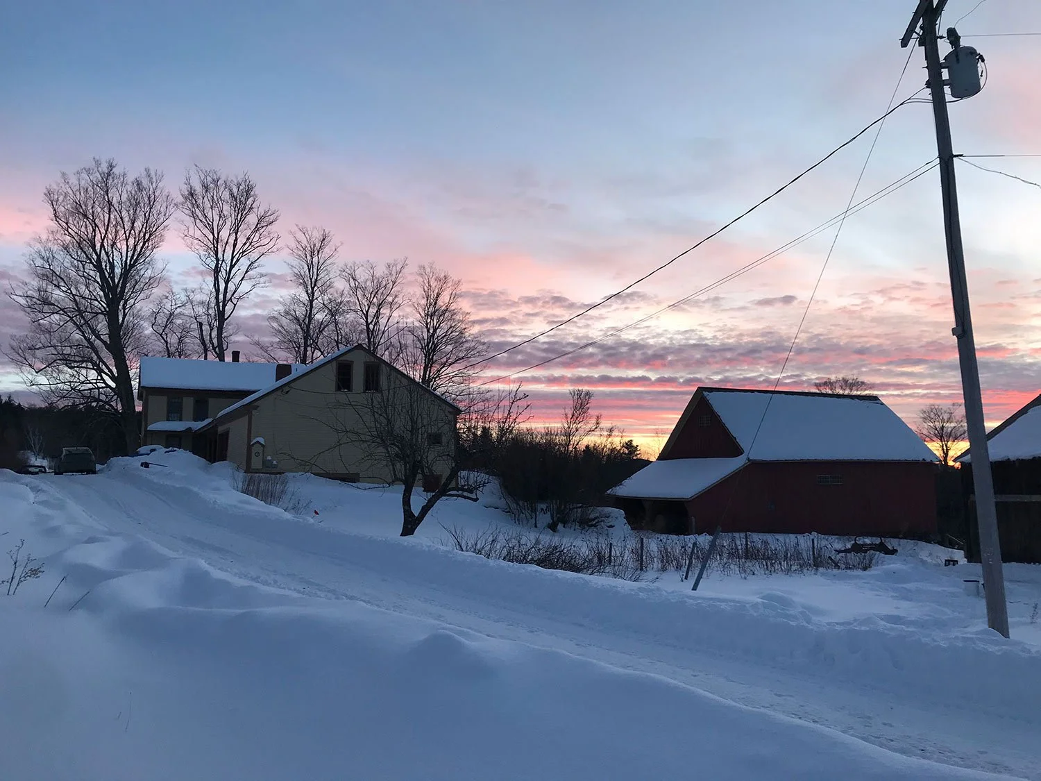 Snow-covered landscape with houses, trees, and power lines at sunset, displaying pink clouds in the sky.