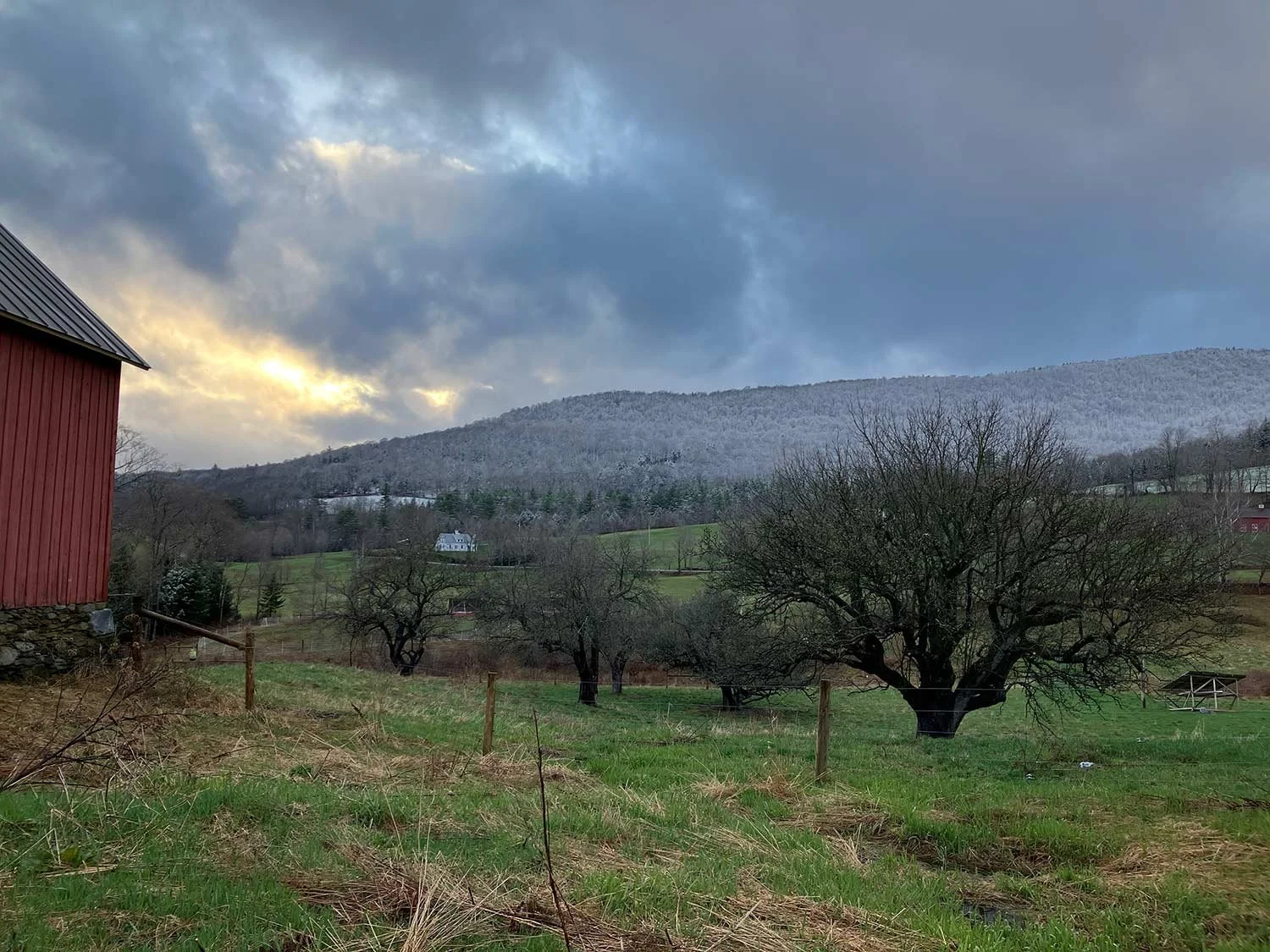 A rural landscape with a red barn on the left, leafless trees in the foreground, green grassy fields, and a mountain range under a cloudy sky.