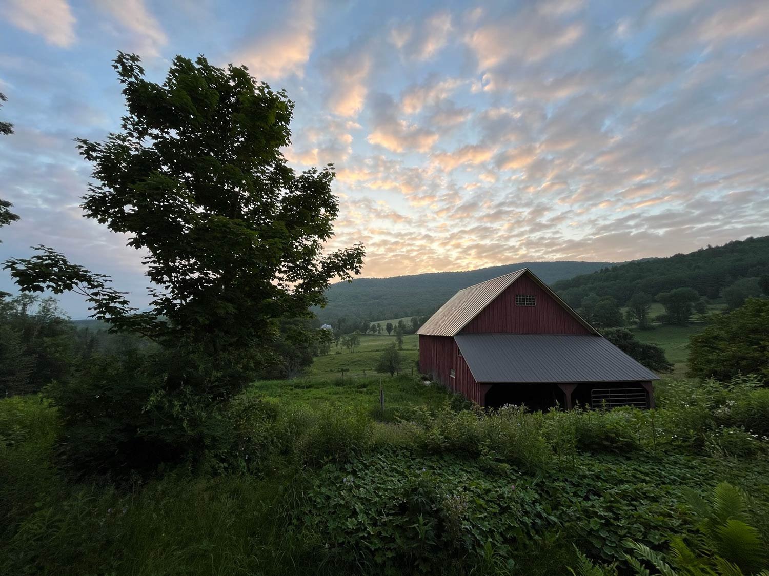 A rural landscape at sunset with a red barn, green trees, rolling hills, and a partly cloudy sky.