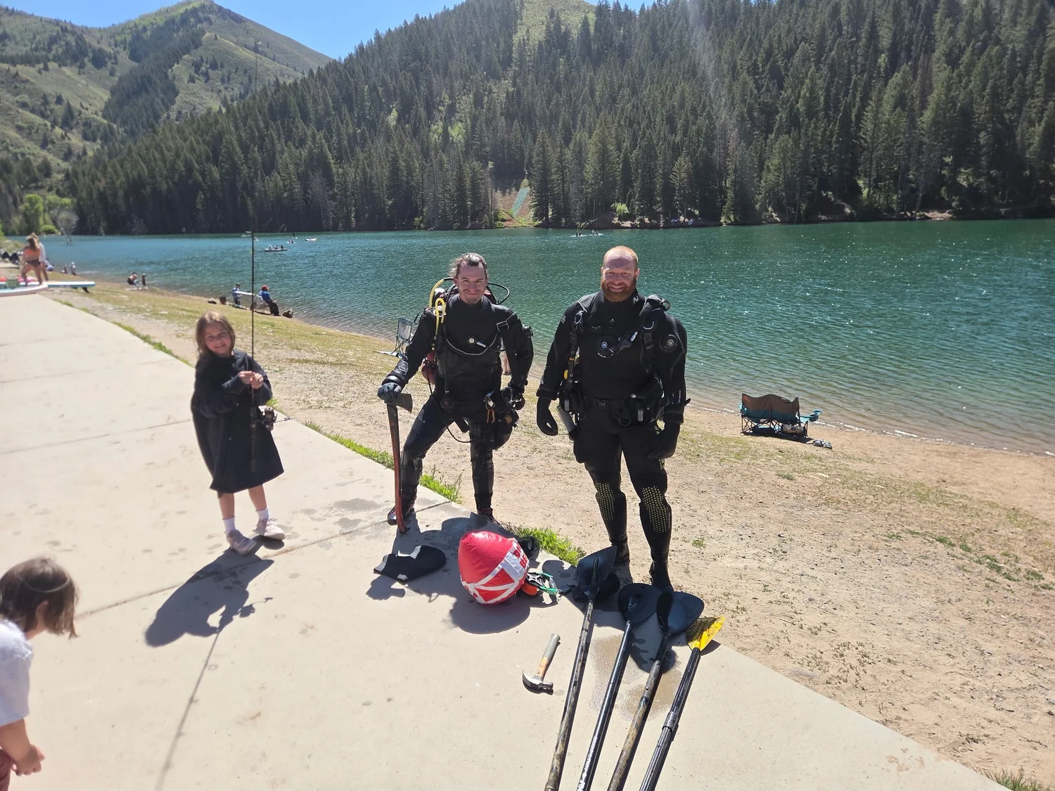 Two Fathom Restoration divers in drysuits standing with fins and gear on the Tibble Fork shoreline, the reservoir and the canyon behind them