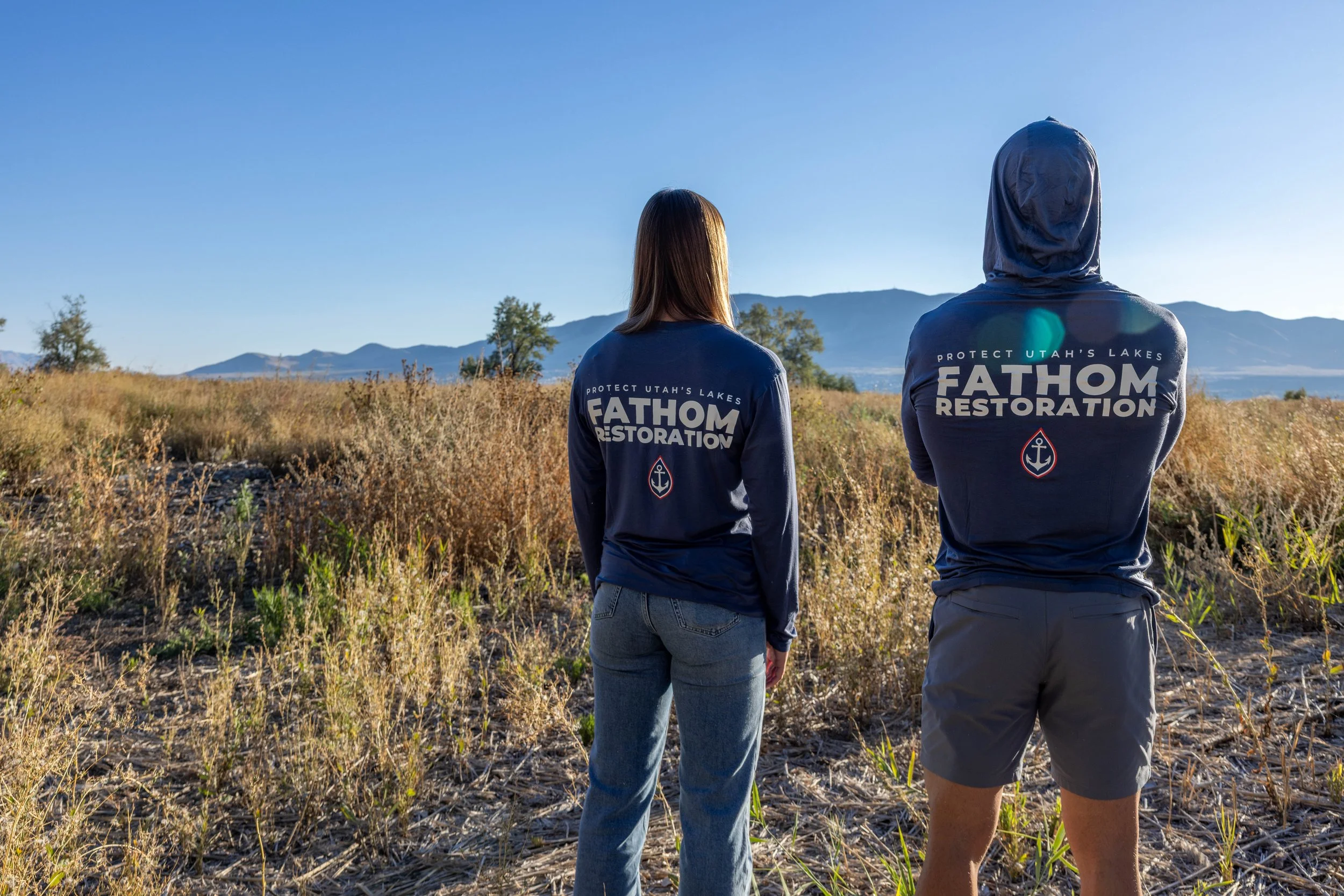 Two people in Protect Utah's Lakes shirts, backs to the camera, facing Utah Lake and the mountains