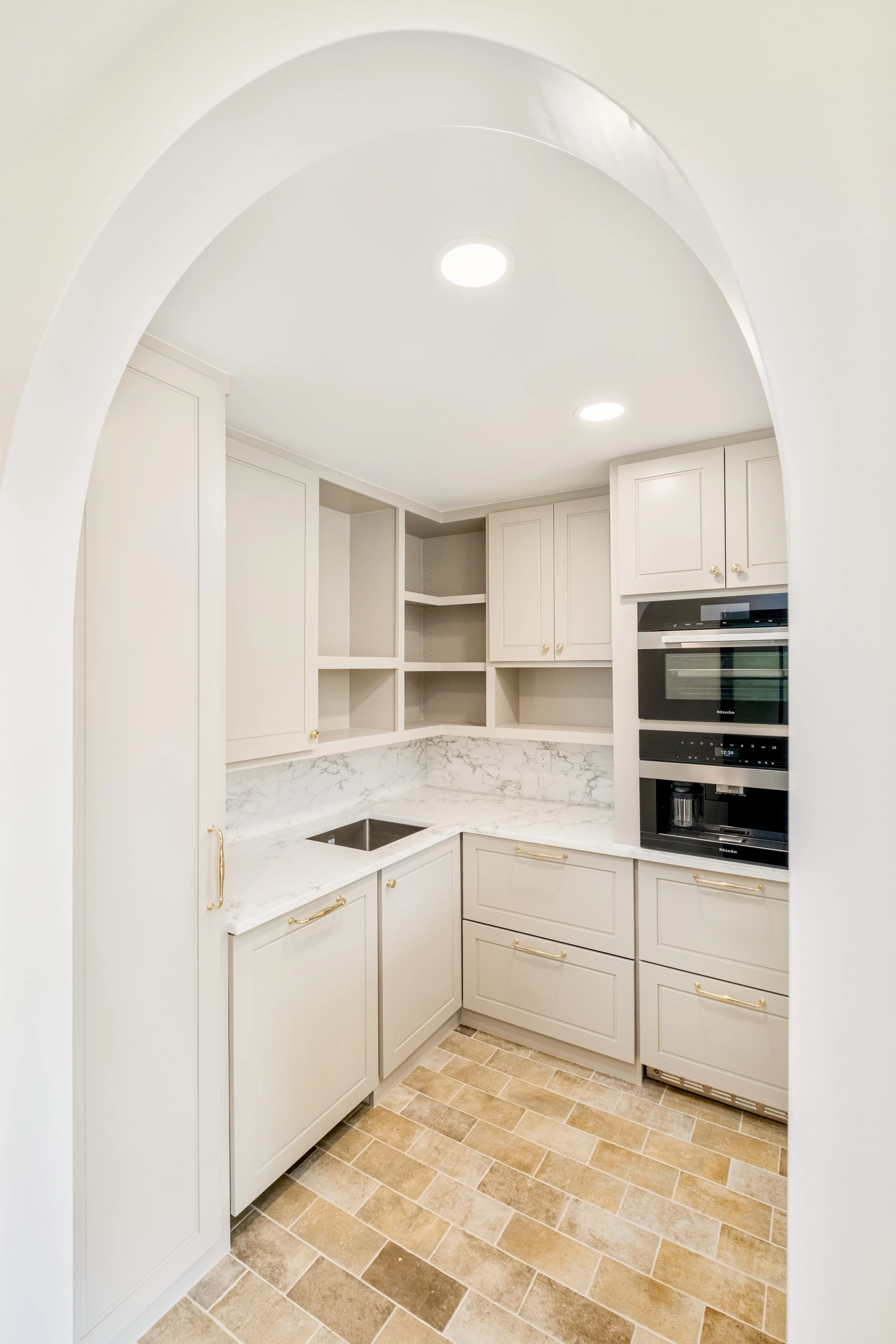 White kitchen with white cabinets, marble countertop, built-in oven, and tiled floor, viewed through an arched doorway.