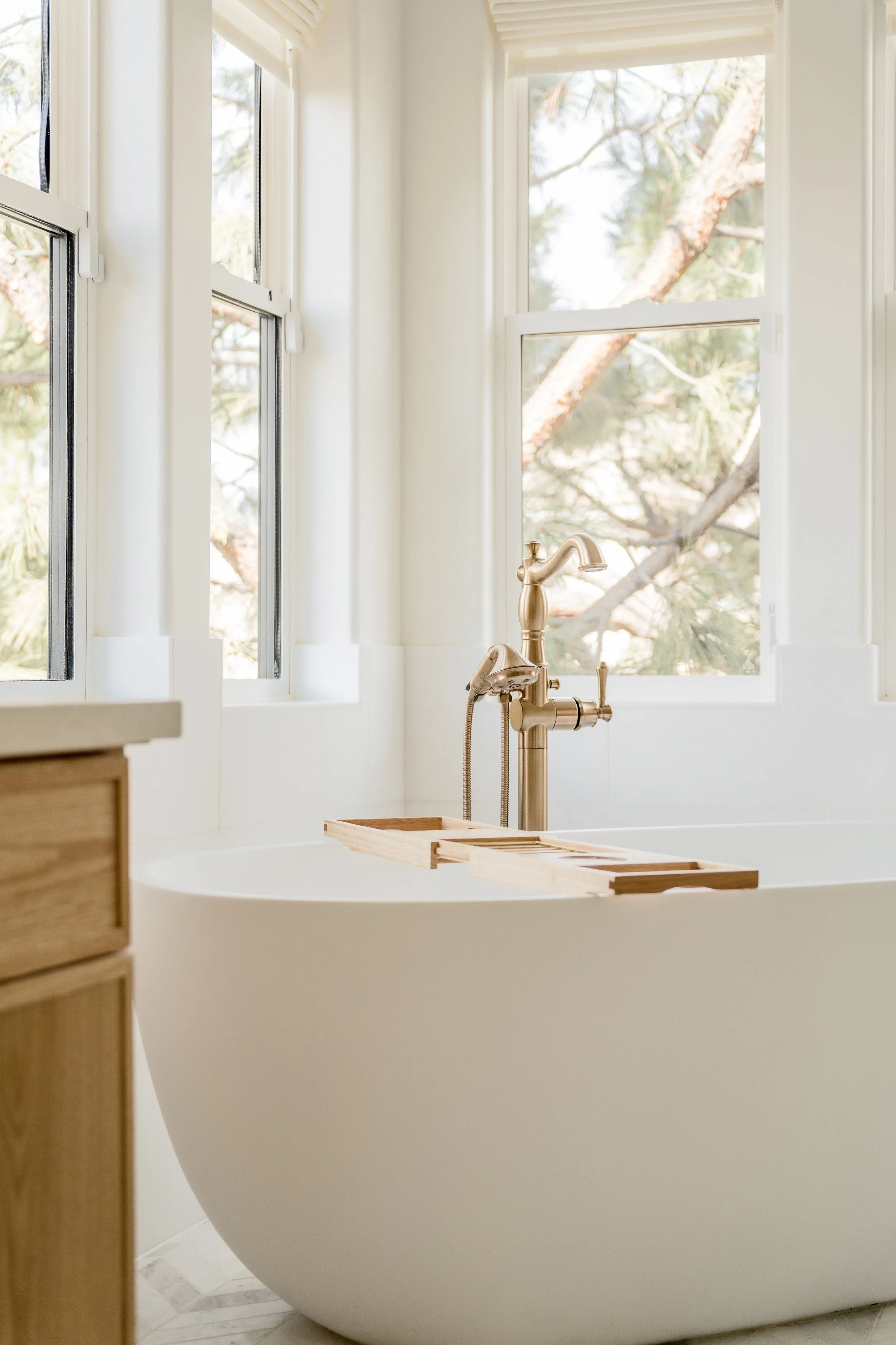 A white bathtub with a gold-colored faucet and a wooden bath tray, set in front of a window with white frames, through which trees are visible outside.