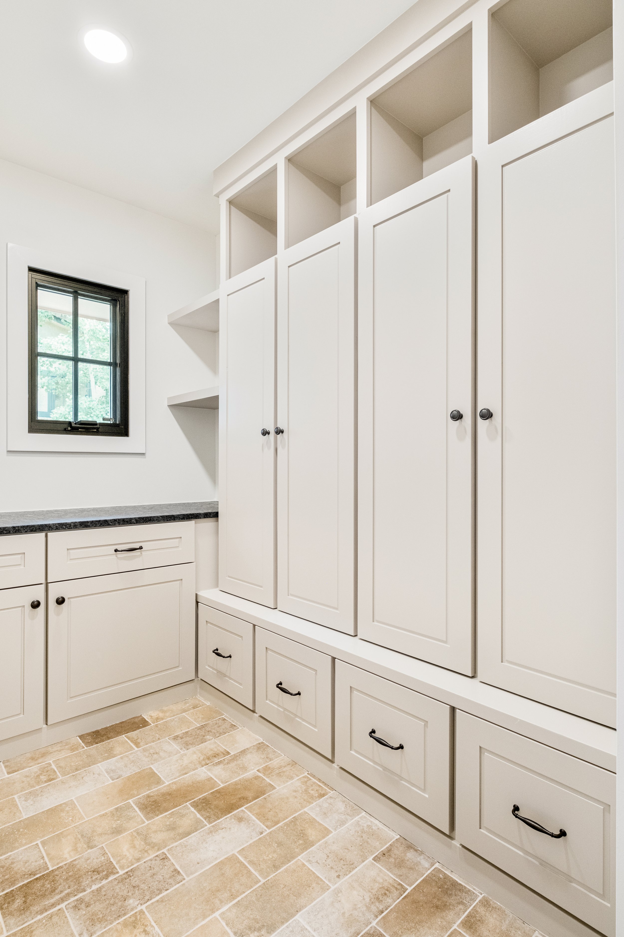 Interior of a laundry room with white cabinets, some with doors and others open shelves, beige tiled floor, and a small window on the left side.