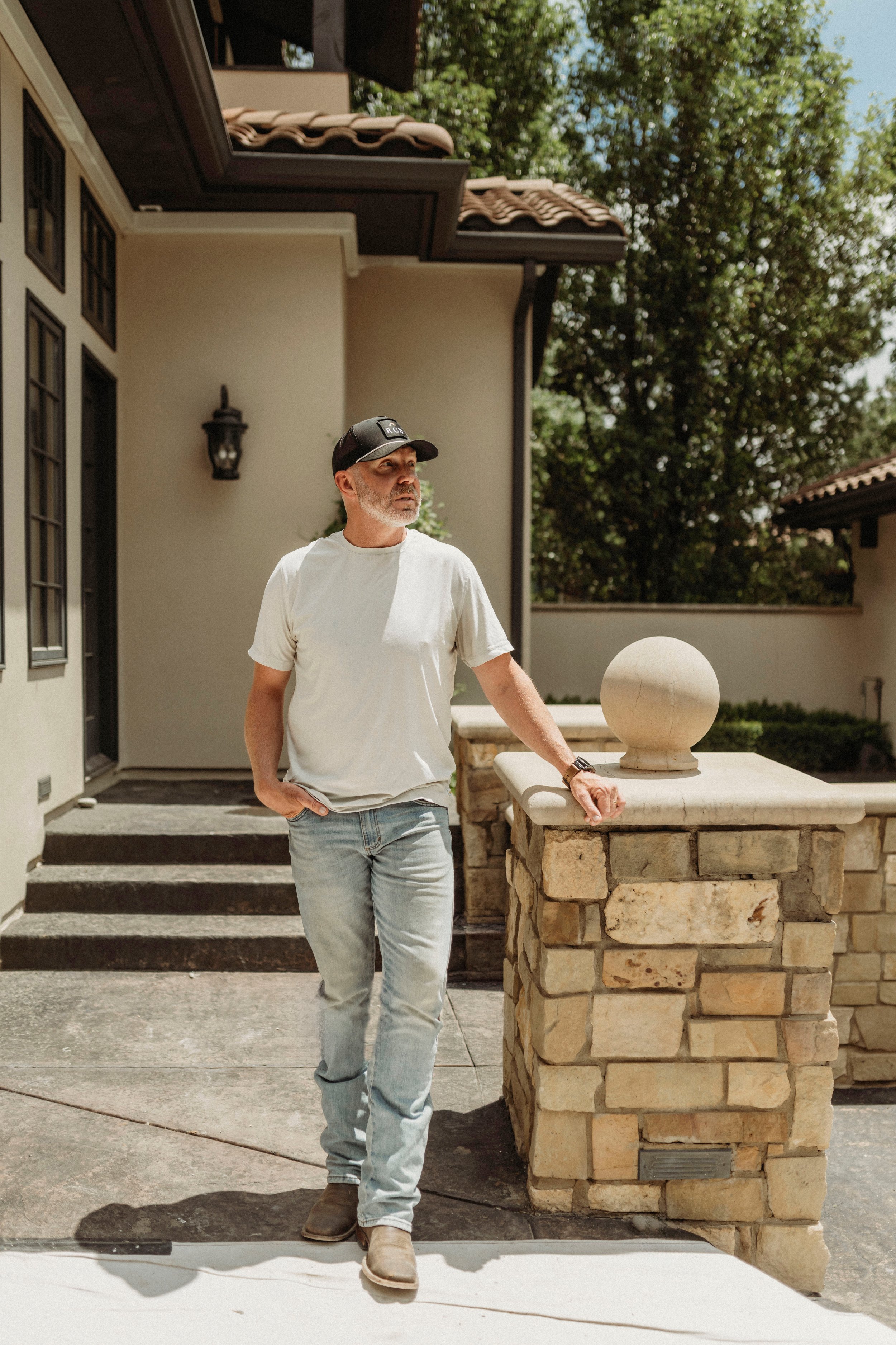 A man with gray beard wearing a black cap, white t-shirt, and jeans, standing on front steps of a house with a stone pillar and trees in the background.
