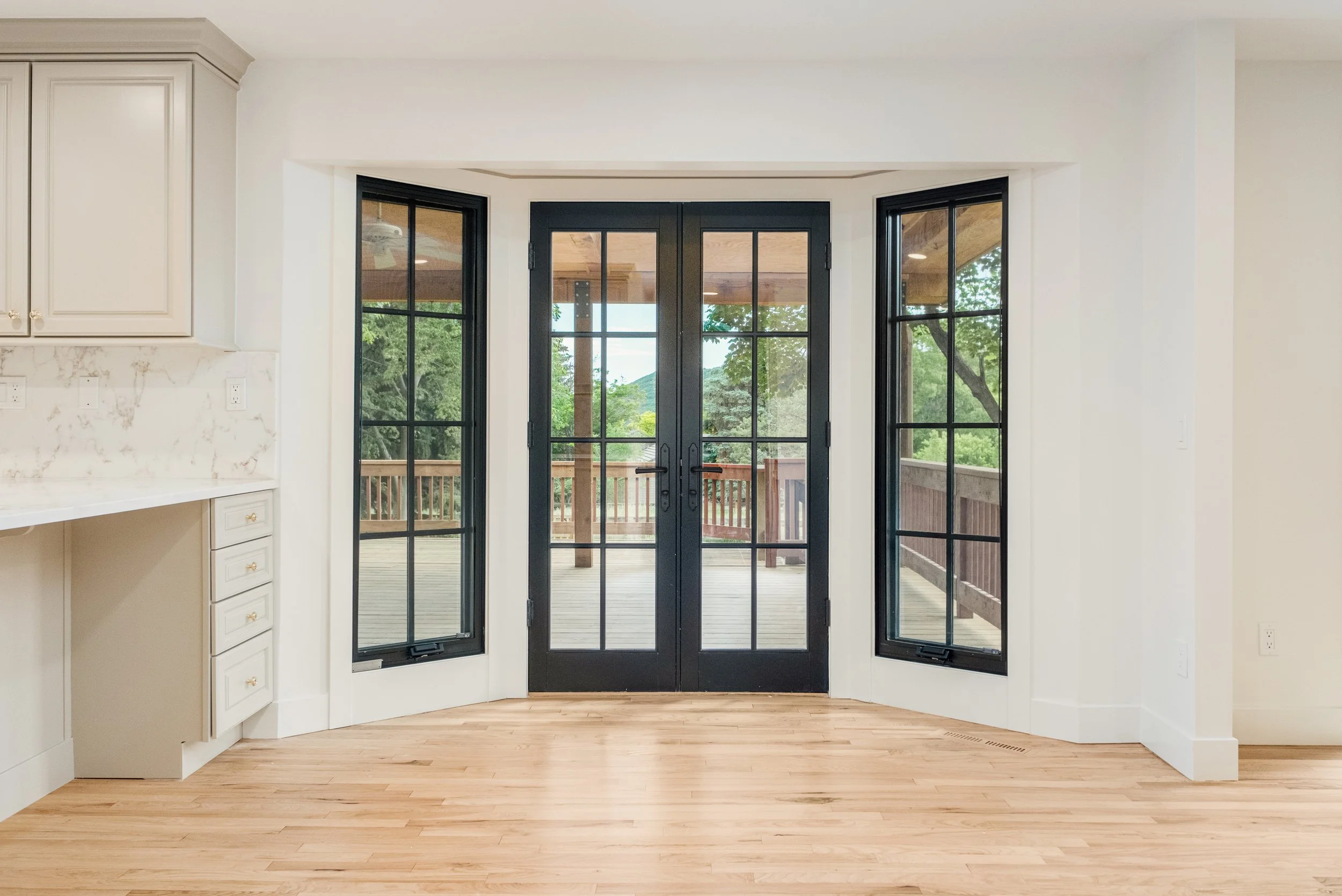 A room with light wood flooring, white walls, and black-framed French doors leading to a deck with a view of green trees outside.