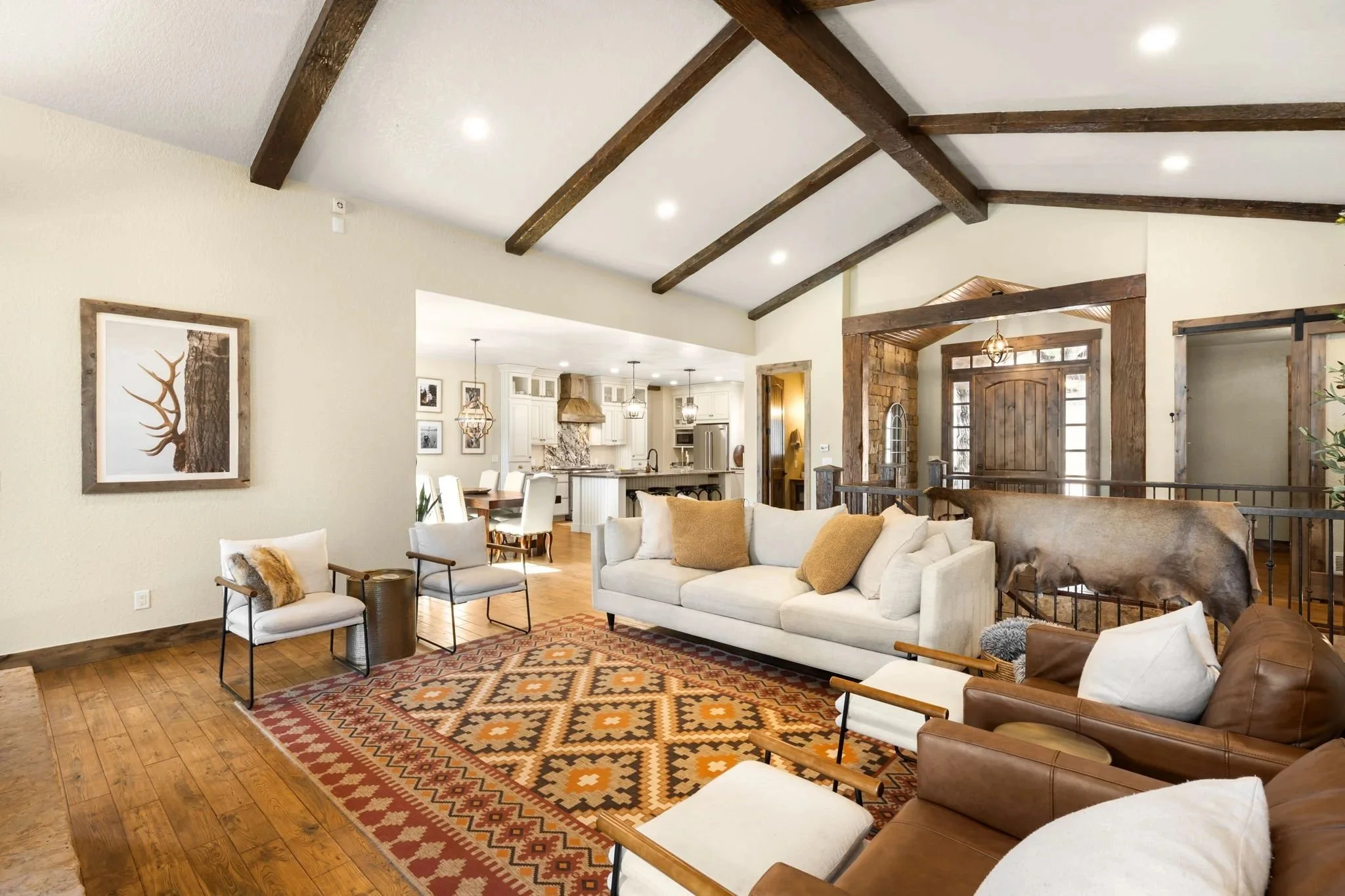 Living room with white sofa, chairs, and a patterned area rug, featuring wood ceiling beams and open kitchen in background.