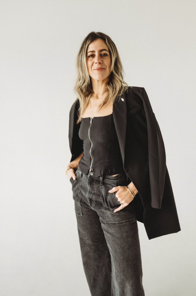 Woman with wavy blonde hair, wearing a black zip-up top, black blazer draped over shoulders, and high-waisted black jeans, standing against a plain white background.