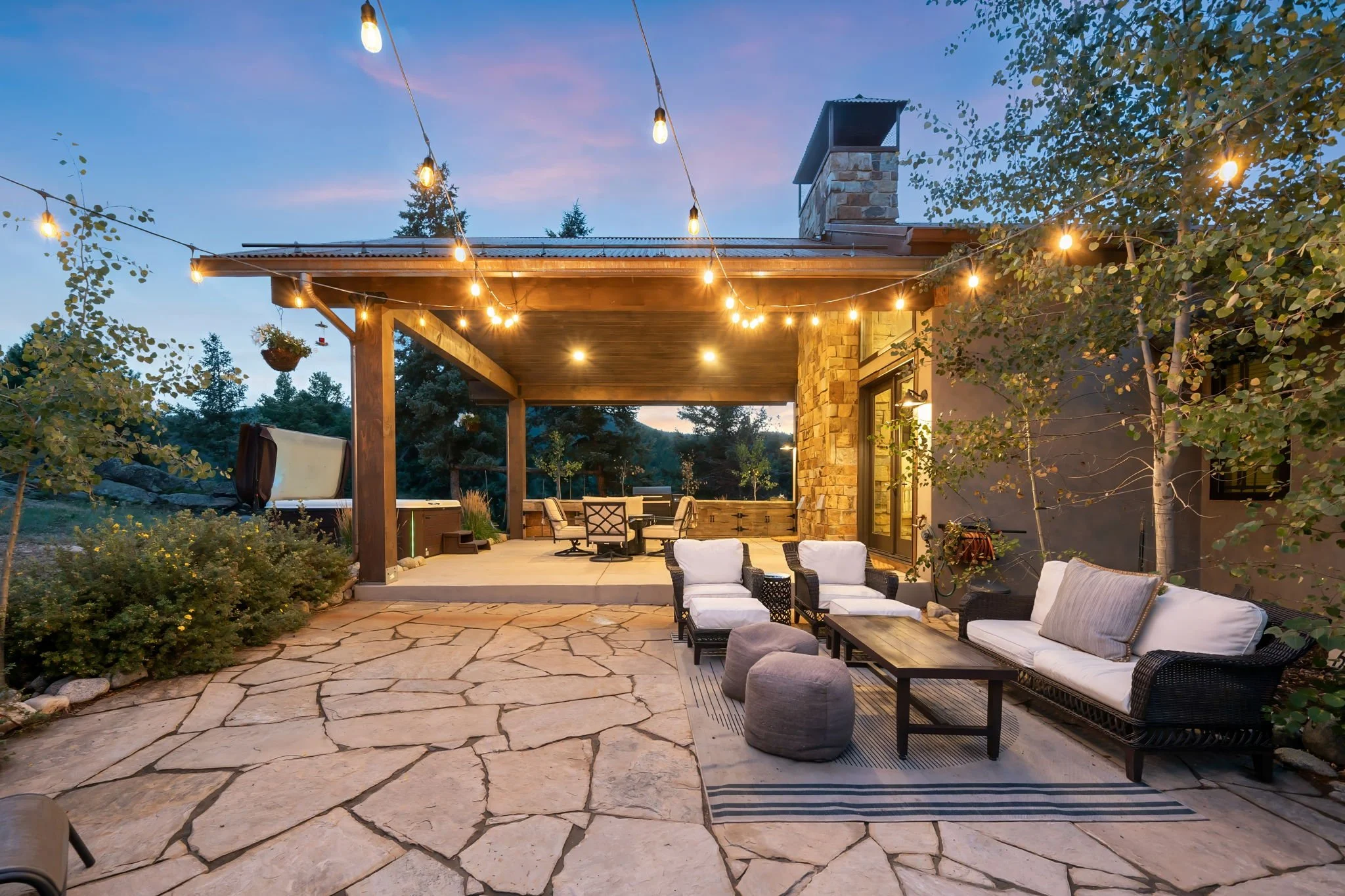 An outdoor patio area with string lights hanging above, surrounded by trees and a house with stone and wood exterior. There are seating arrangements including cushioned chairs, a table, and a lounge chair, with a view of a wooded landscape at dusk.
