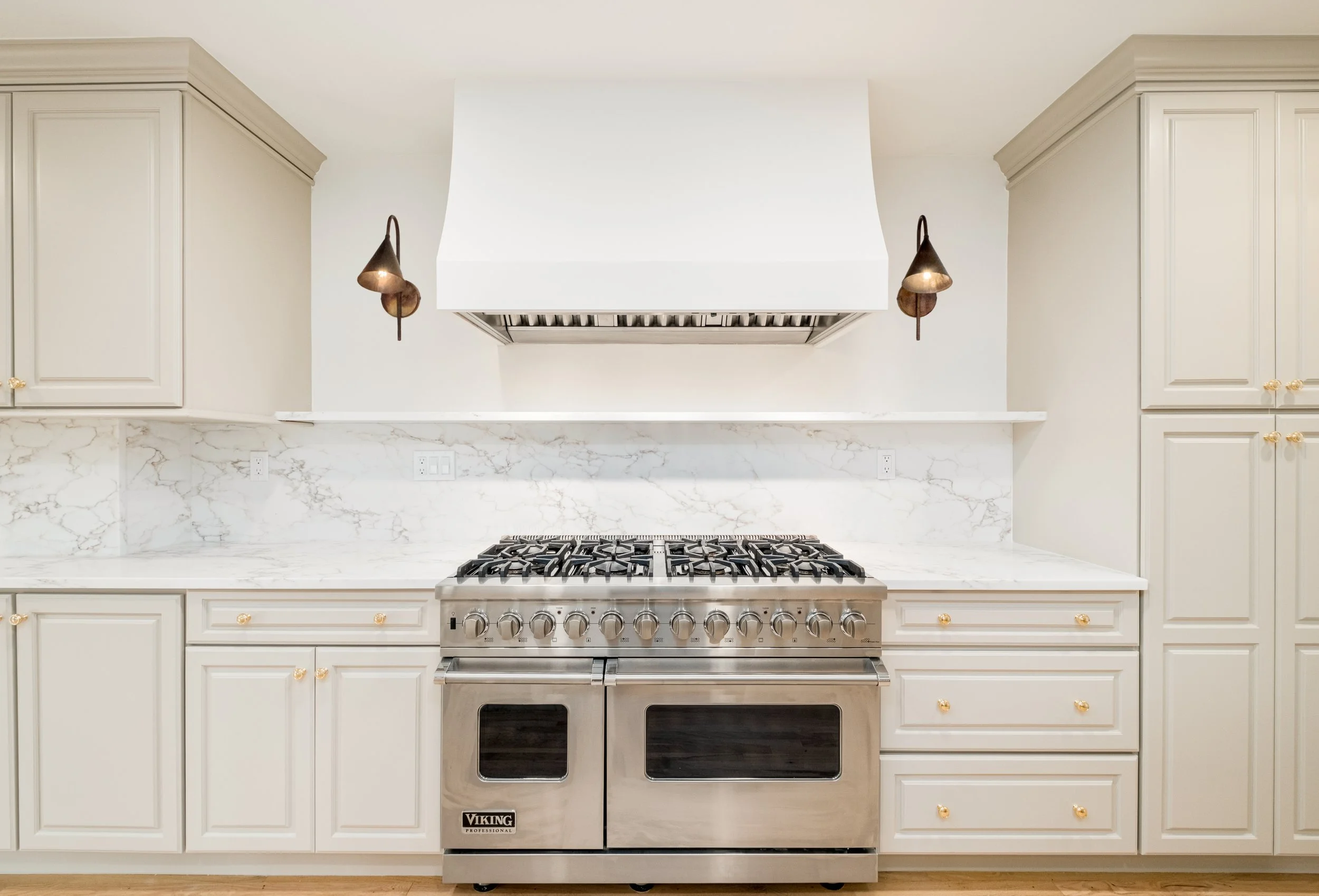 A modern kitchen with white cabinetry, a marble countertop, and a stainless steel Viking professional gas range with oven. There are two brass wall sconces on either side of a white range hood.