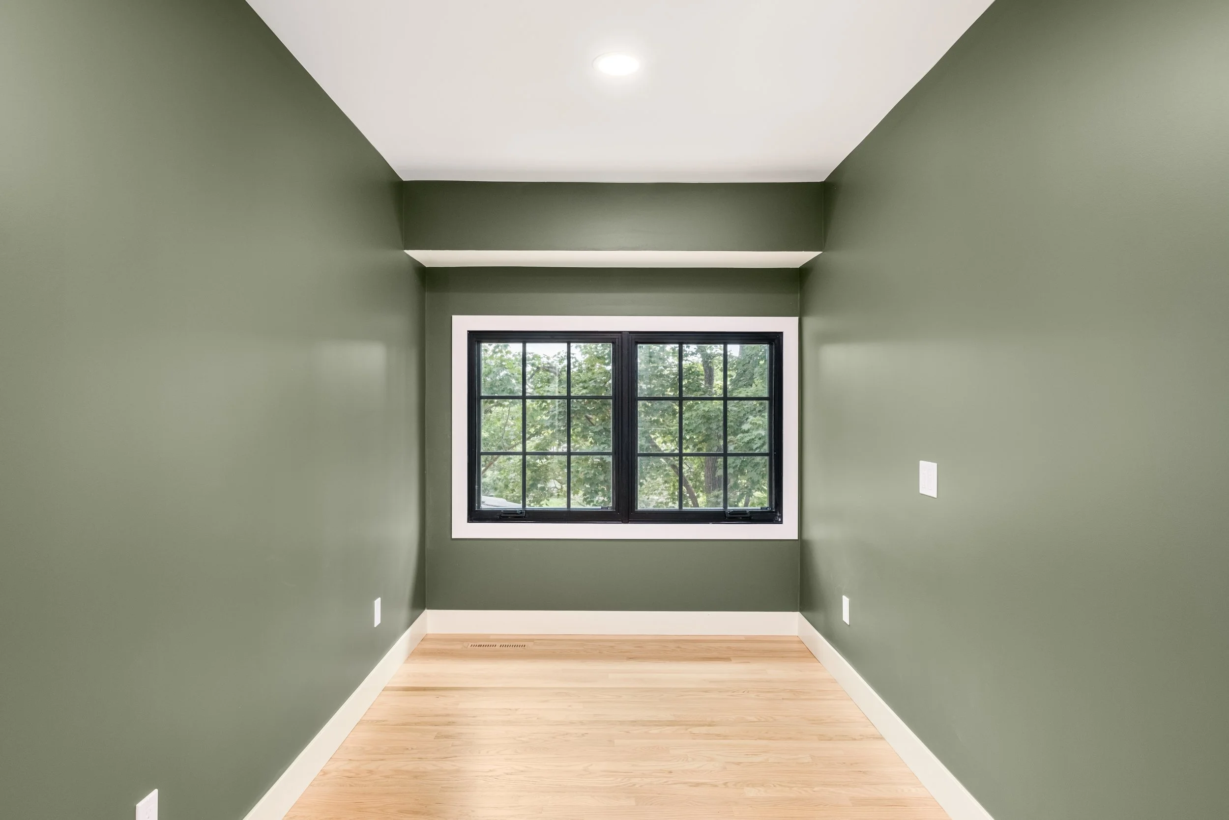 A small, empty room with green walls, a white ceiling, light wood flooring, and a single black-framed window with a view of green trees outside.
