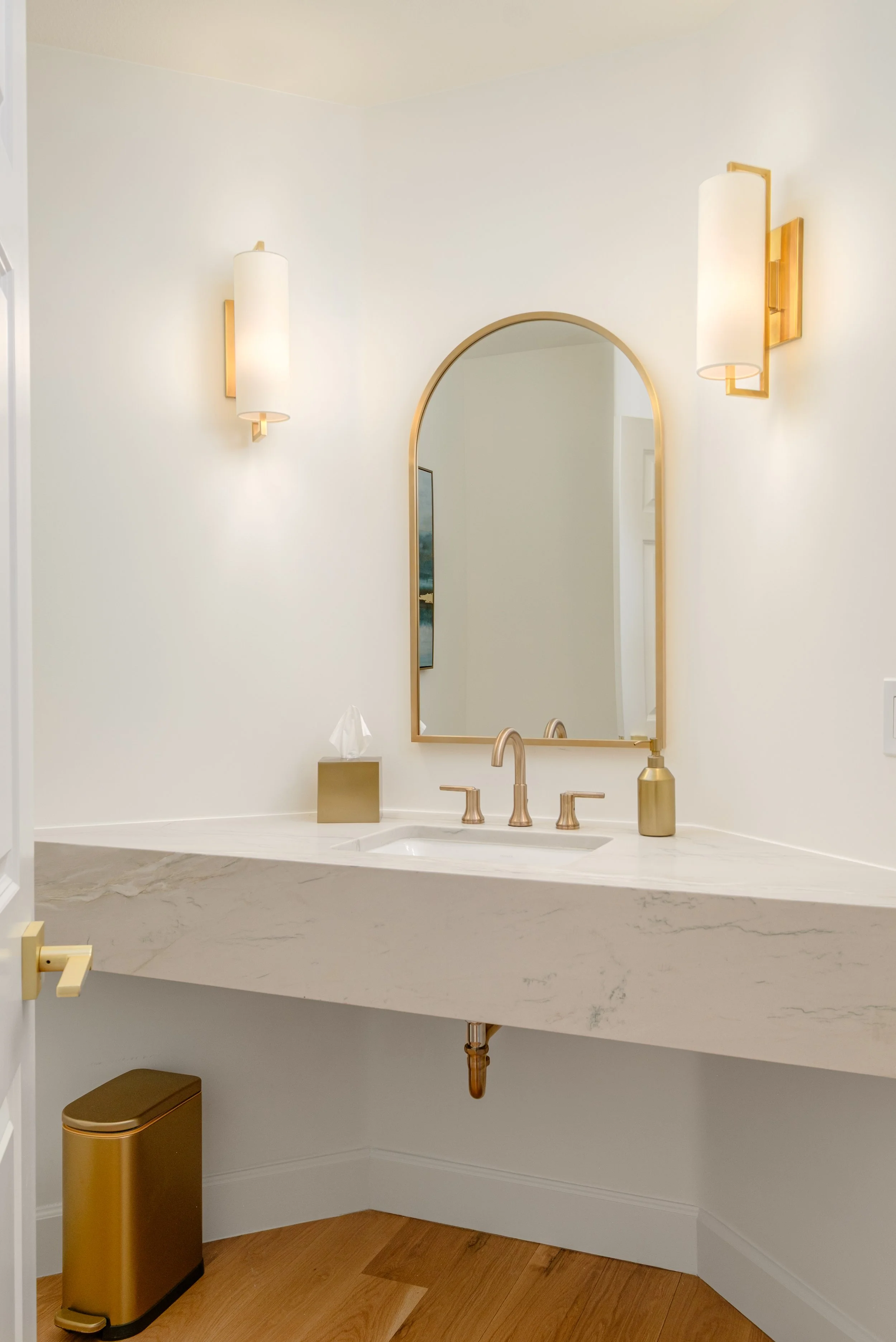 Bathroom vanity with oval mirror, gold fixtures, two white wall sconces, a tissue box, and a gold trash can.