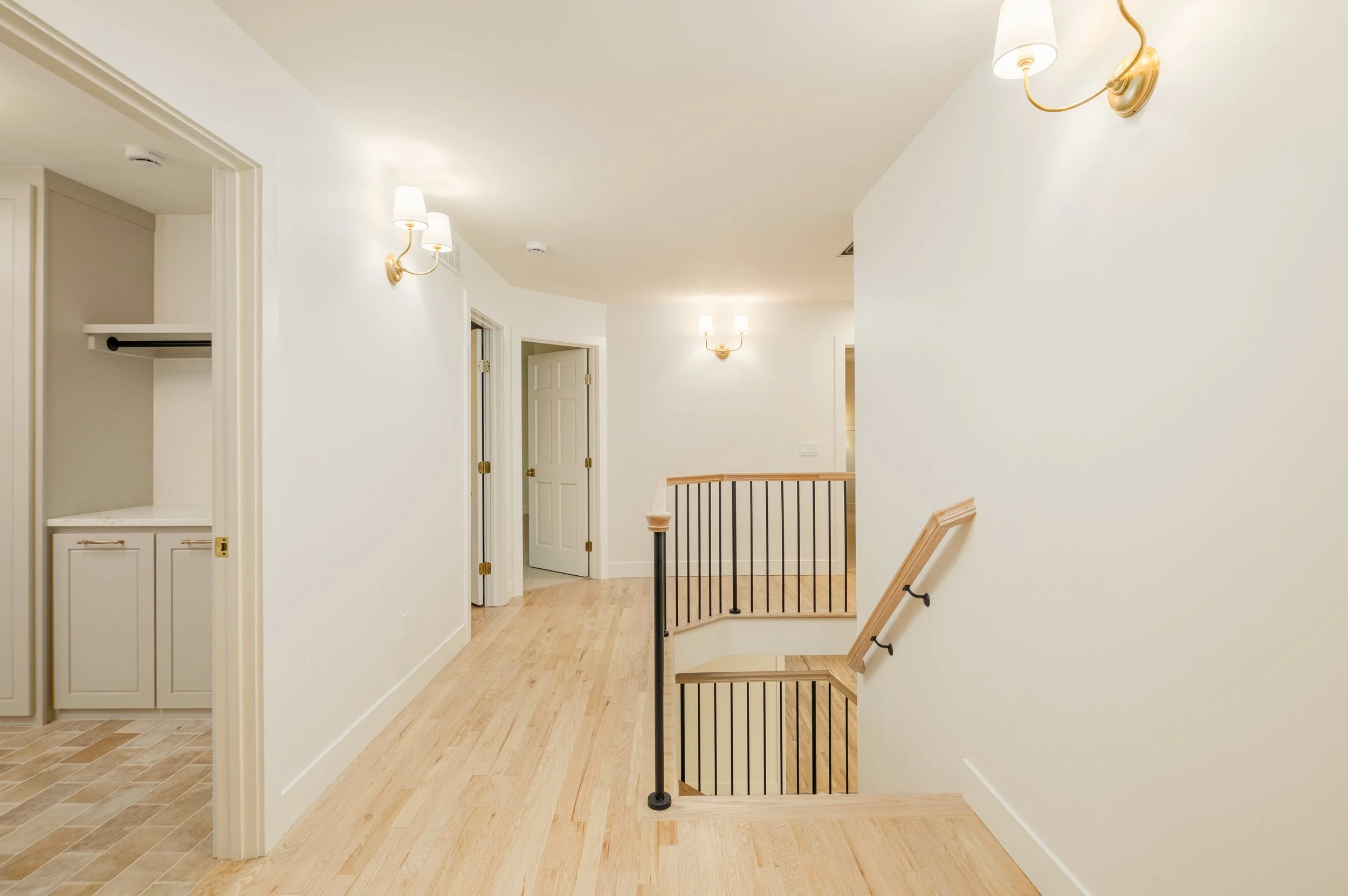Interior view of a house hallway with wooden floors, white walls, wall-mounted sconces, doors, and an open staircase with a wooden handrail and black metal balusters.