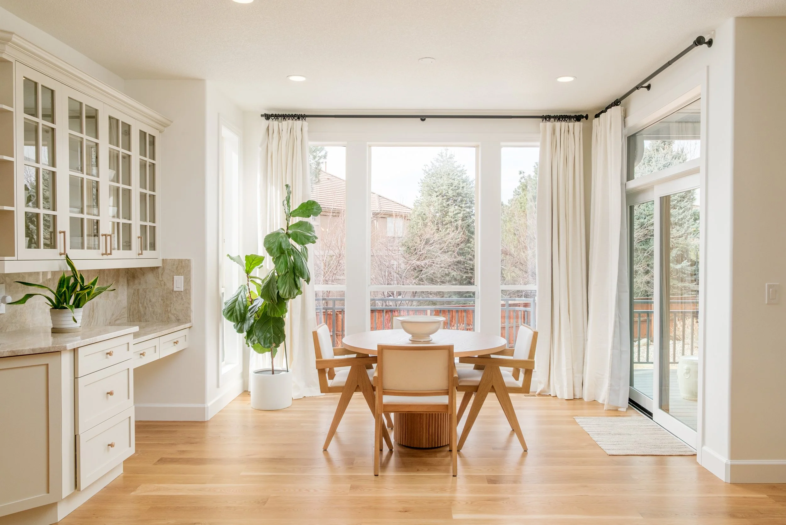 A bright dining area with large windows, white curtains, a round wooden table with four chairs, and a potted plant in a white container.