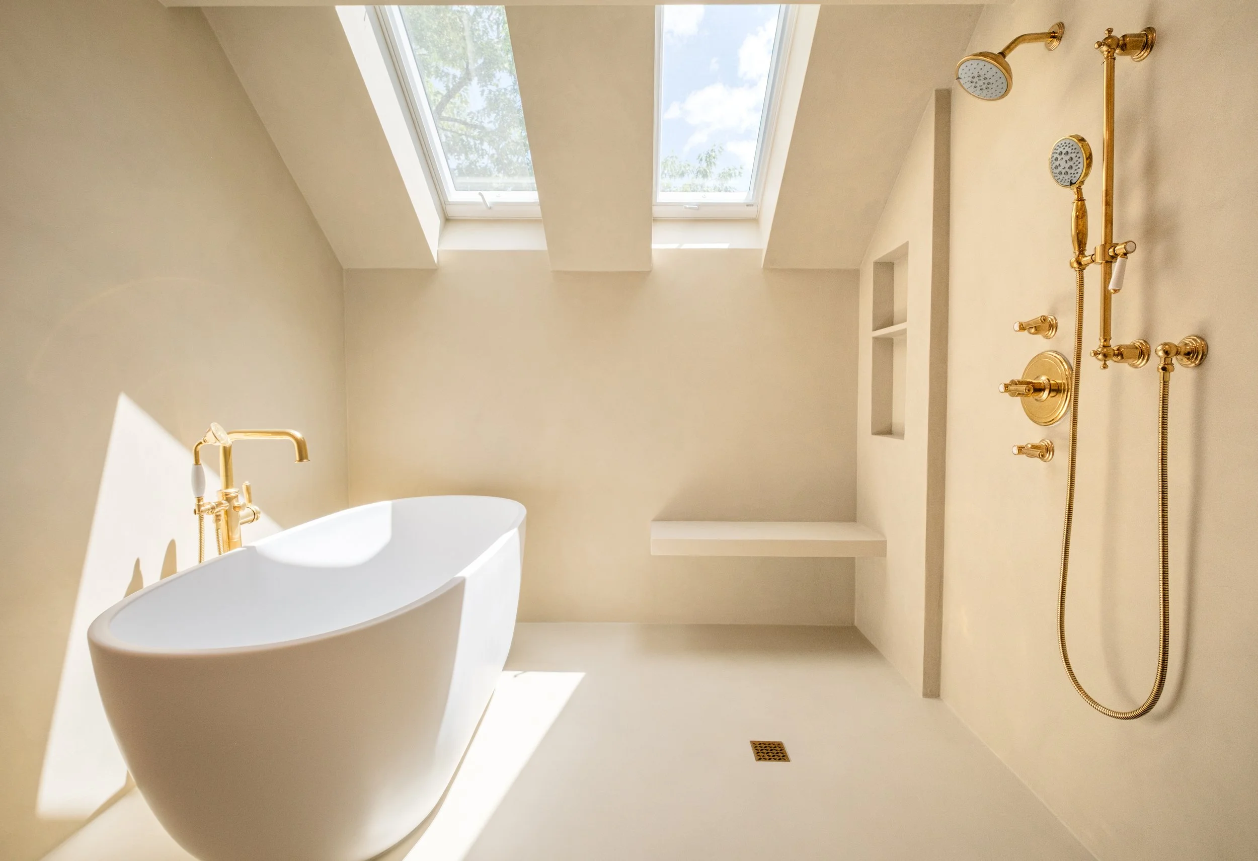 A bathroom with a skylight window, a white free-standing bathtub, and gold fixtures including a showerhead, hand shower, and faucet. The walls are painted a light color, and there are small built-in shelves in the wall.