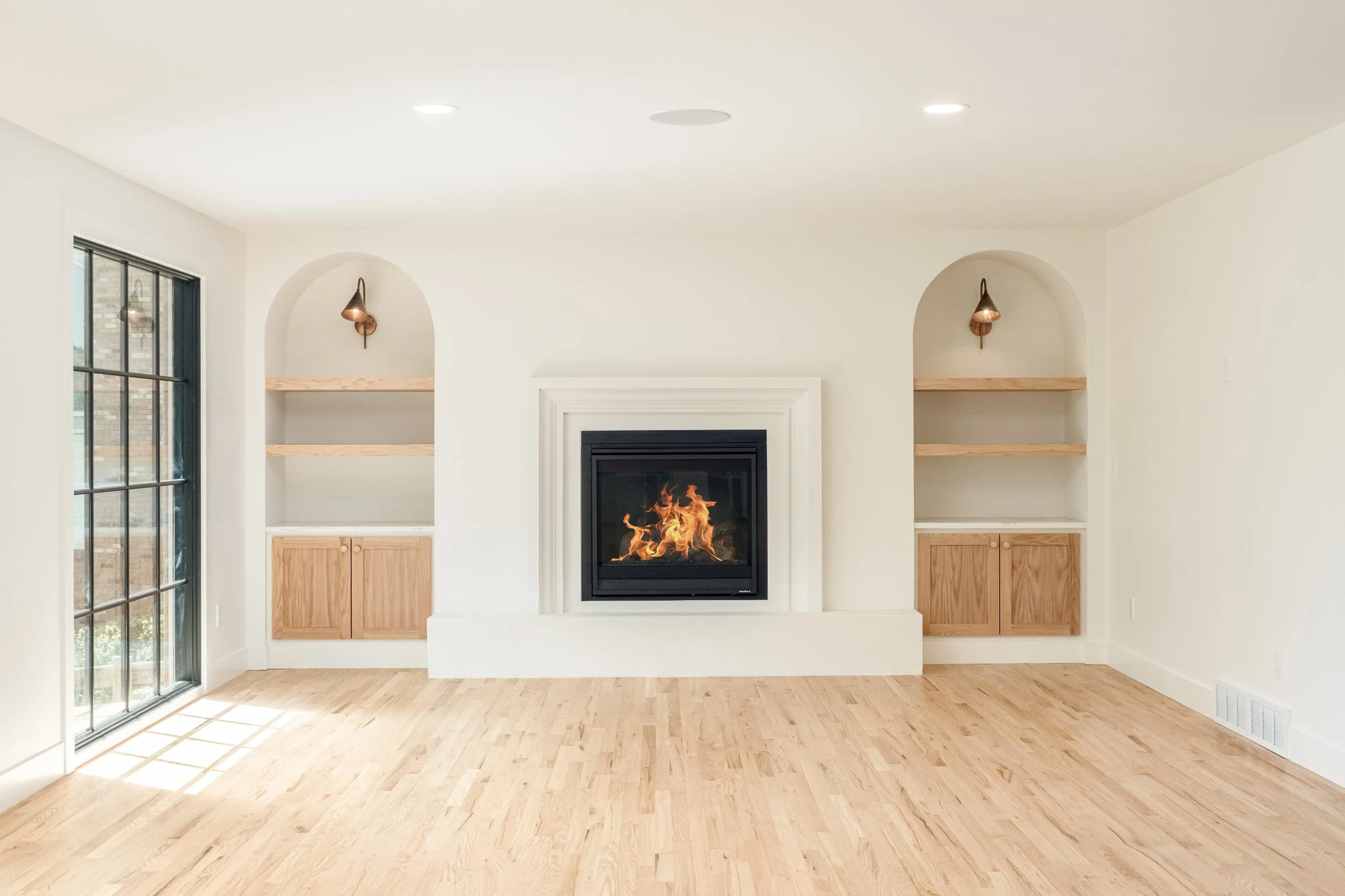 Living room with a fireplace, light wood flooring, two wooden built-in shelves on each side of the fireplace, large window on the left, and recessed ceiling lights.