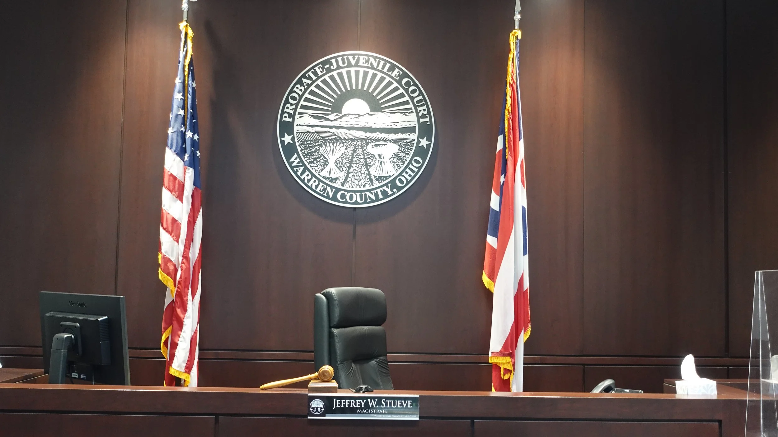 A courtroom setting featuring a dark wood panel backdrop with a circular emblem that reads 'Probate-Juvenile Court Warren County, Ohio.' Two American flags are positioned on either side of the emblem.