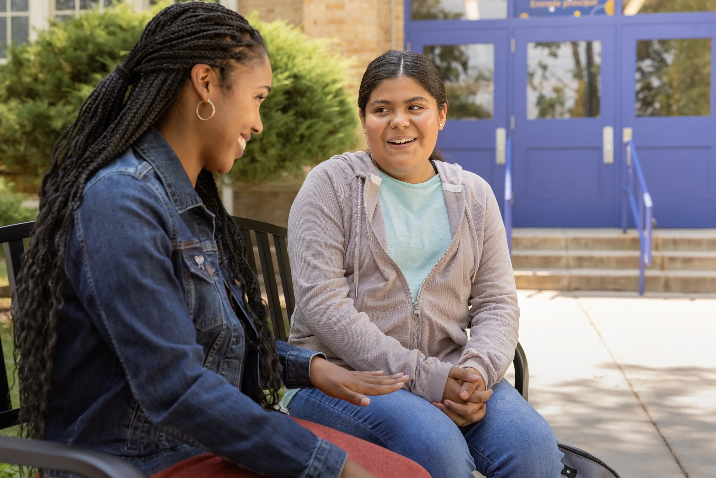 A woman and a child sit outside on a bench, engaged in conversation, with a building and greenery in the background.