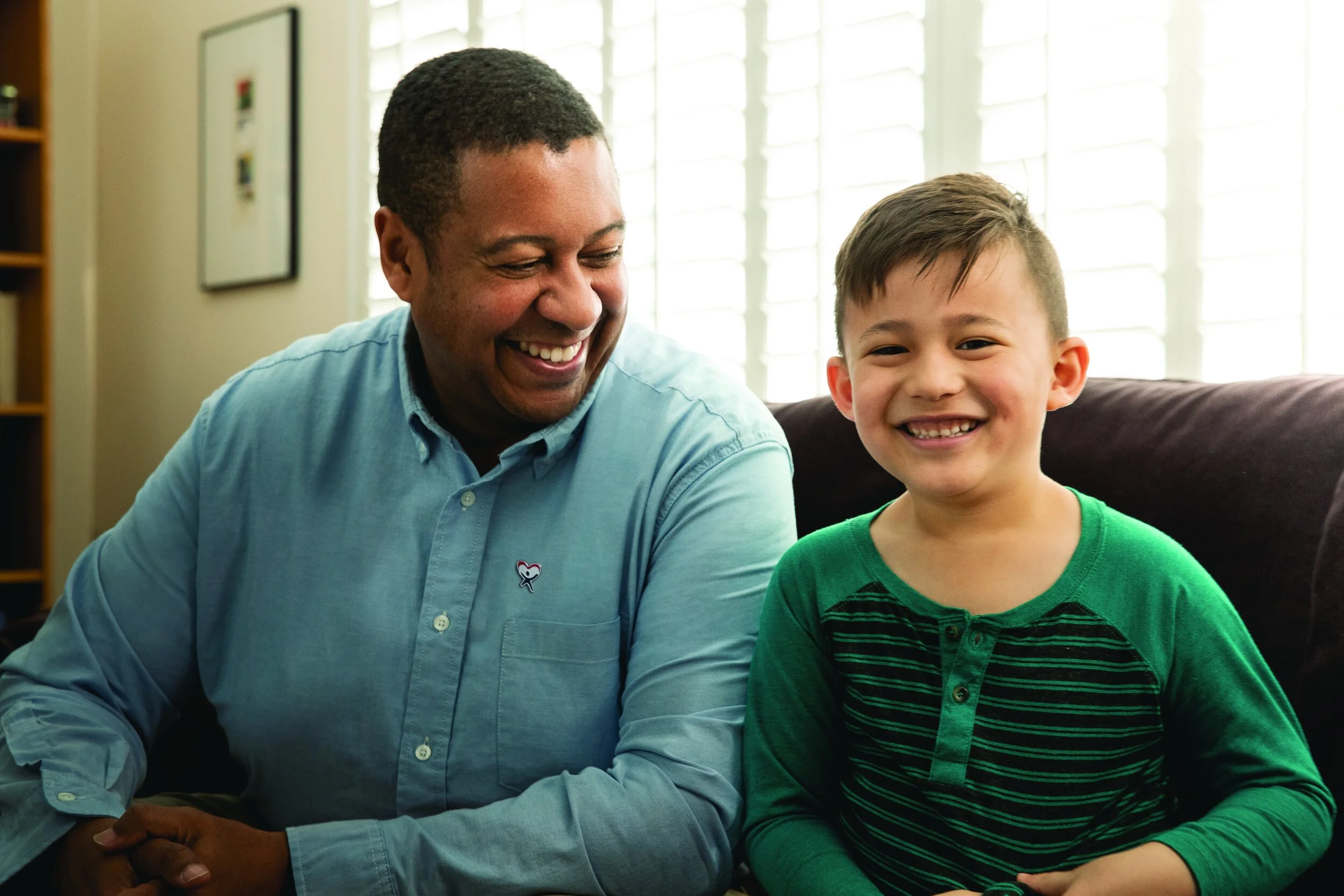 A man and a boy sitting close together indoors next to a window with white blinds, smiling and looking at each other, with the man wearing a light blue shirt and the boy in a green and black striped shirt.
