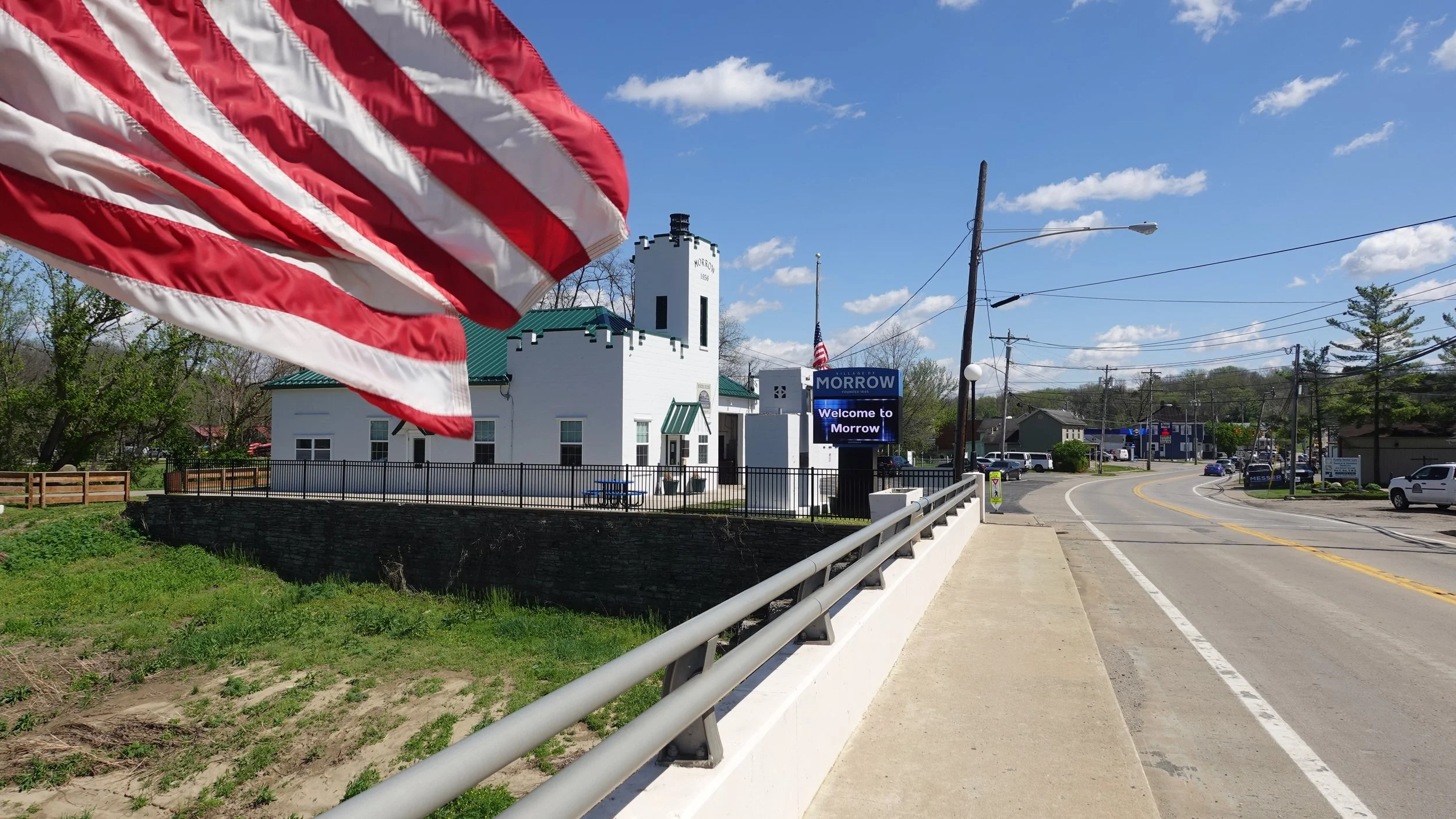 A small town street scene with a white building labeled Morrow and a digital sign welcoming visitors. An American flag is visible in the foreground, partially obscuring the view. There are cars on the road and trees lining the street under a blue sky with some clouds.