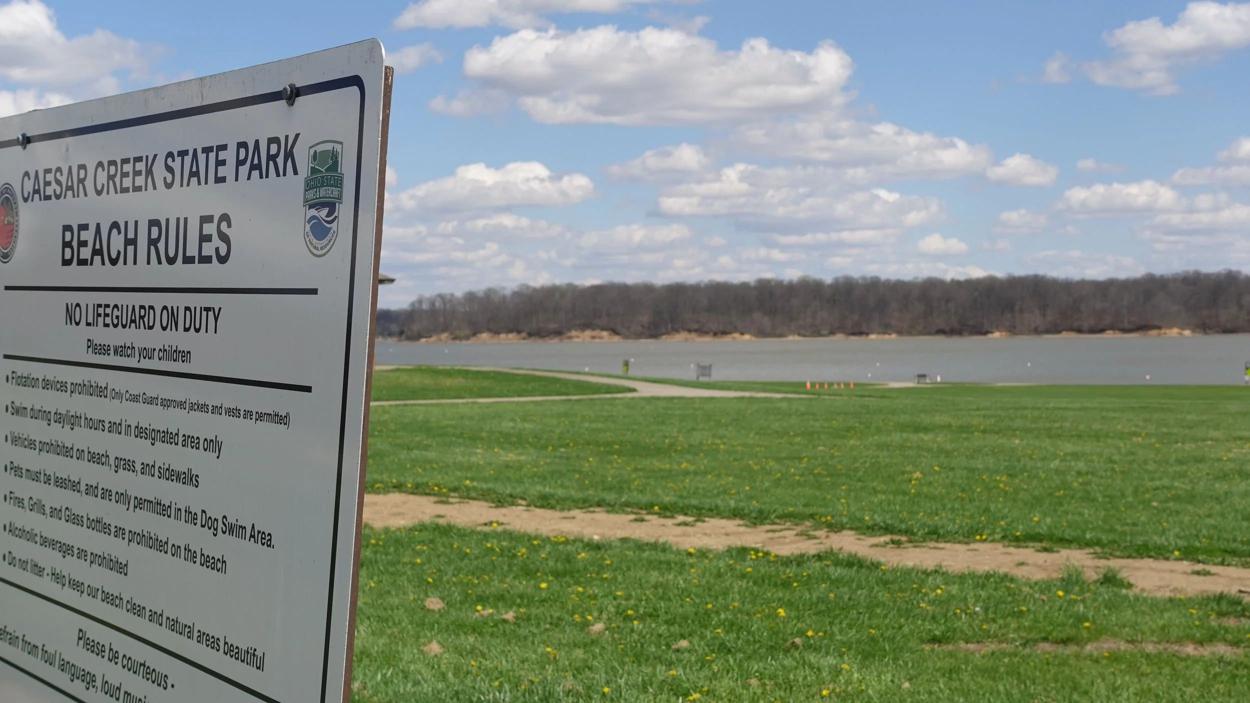 Signboard at Caesar Creek State Park with beach rules, with a view of a lake, green grass, and a blue sky with scattered clouds.