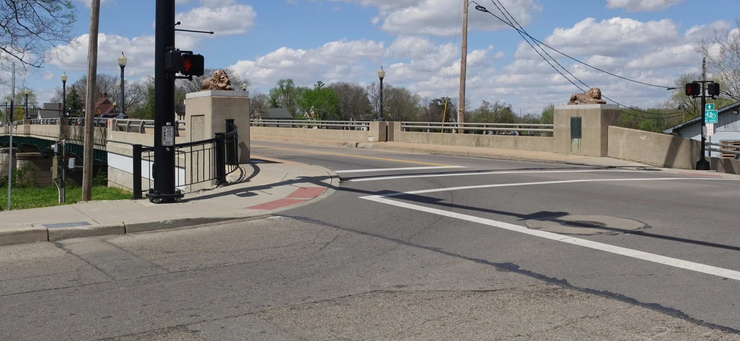 A street crossing with a traffic light, decorative lion statues on concrete pedestals, a bridge in the background, and a bike route sign on a pole under a partly cloudy sky.