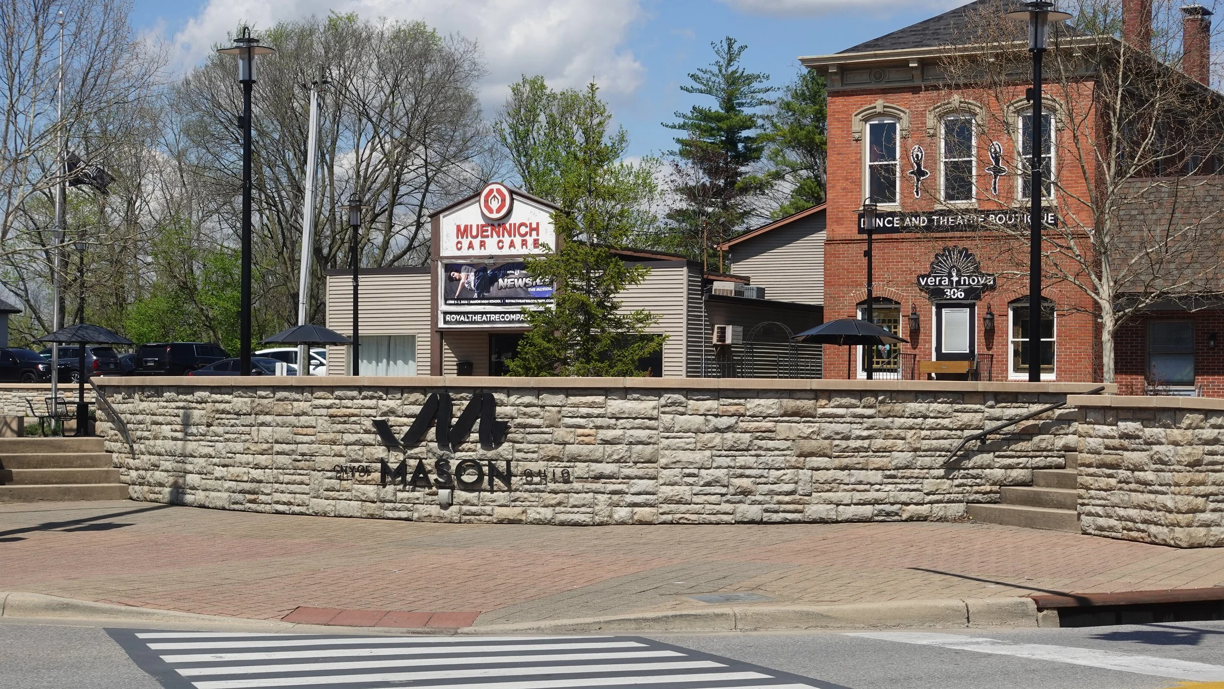 A city of Mason sign with steps leading up to it, surrounded by a stone wall, with street crossing lines visible in the foreground. In the background, there are trees, buildings, and parking lot with cars. The scene is outdoors on a partly cloudy day.