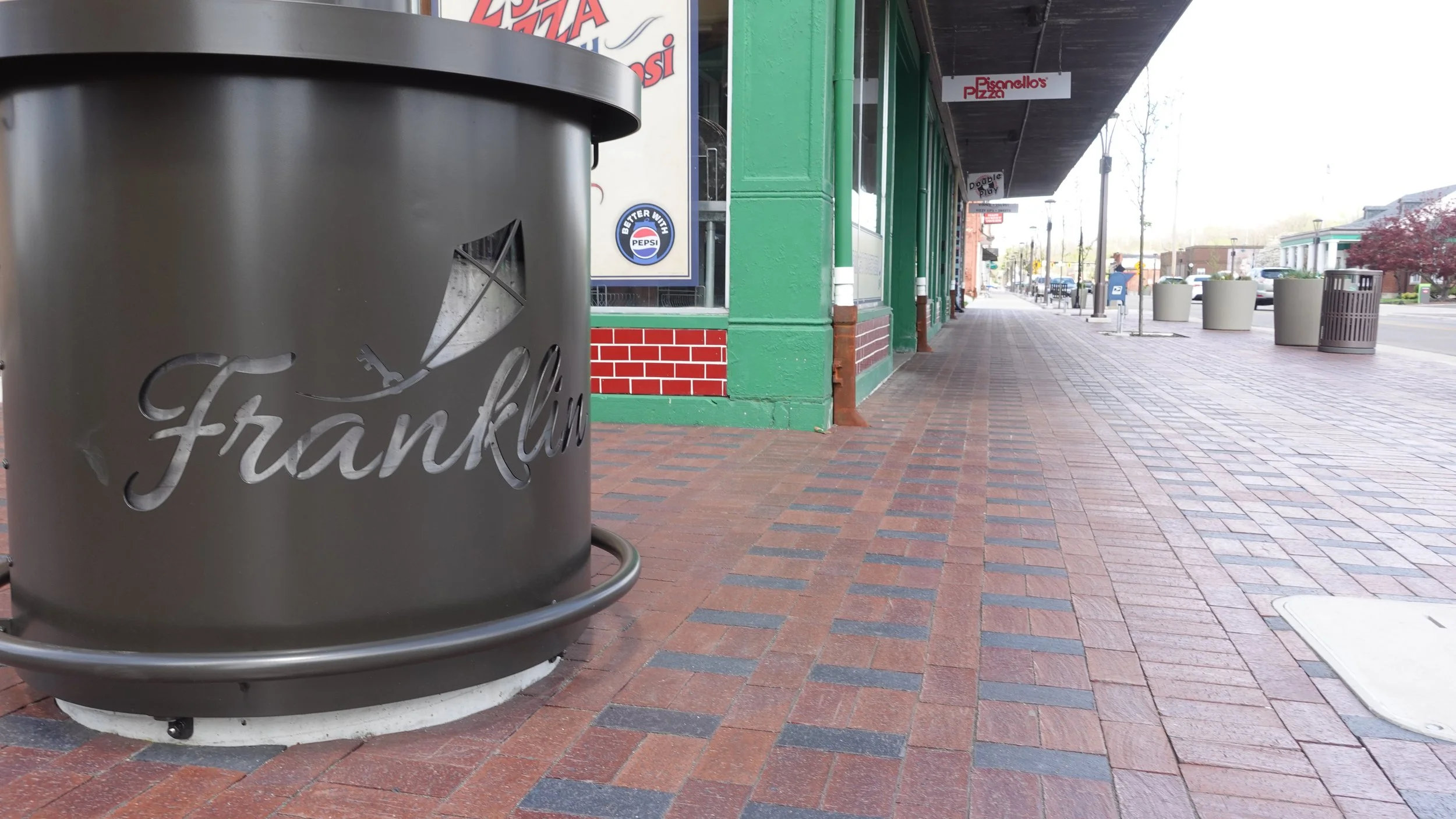 A sidewalk scene featuring a large metal can with the word "Franklin" written on it, adjacent to a green building with brick accents. There are other trash cans, a streetlamp, and storefront signs in the background.